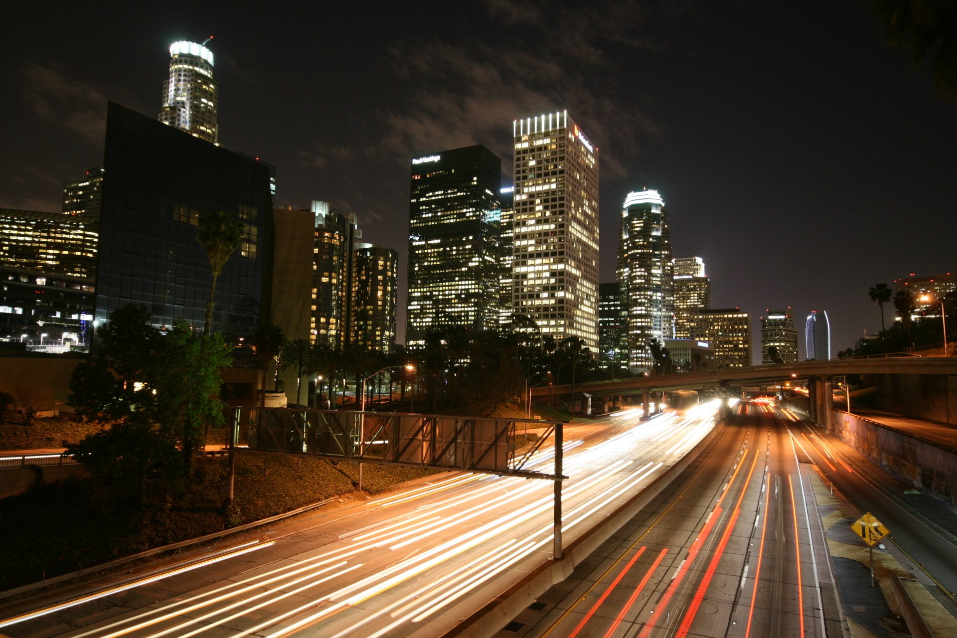Nighttime cityscape with illuminated man-made skyscrapers and a busy road showing streaks of vehicle lights.