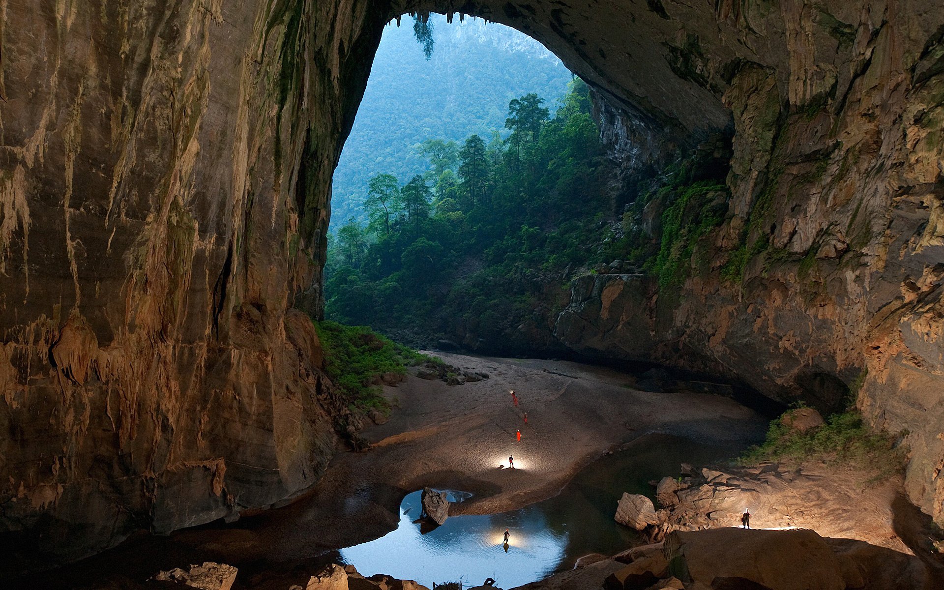 A vast cave entrance opens to a lush, green forested area in nature, with a small river flowing through the rocky cavern floor.
