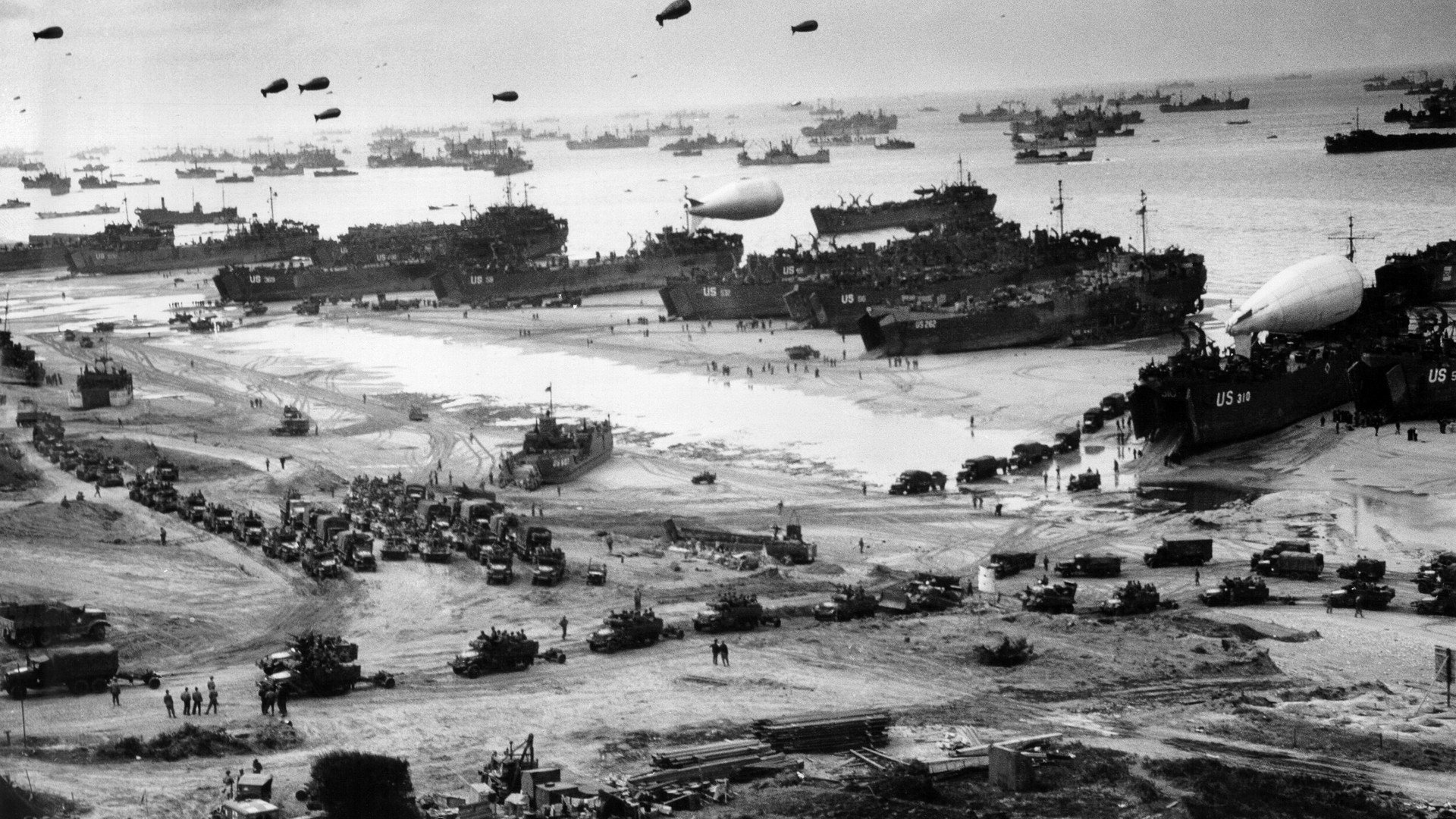Black & white photography capturing a vast military beach landing with numerous ships, vehicles, and soldiers during a historic wartime operation.