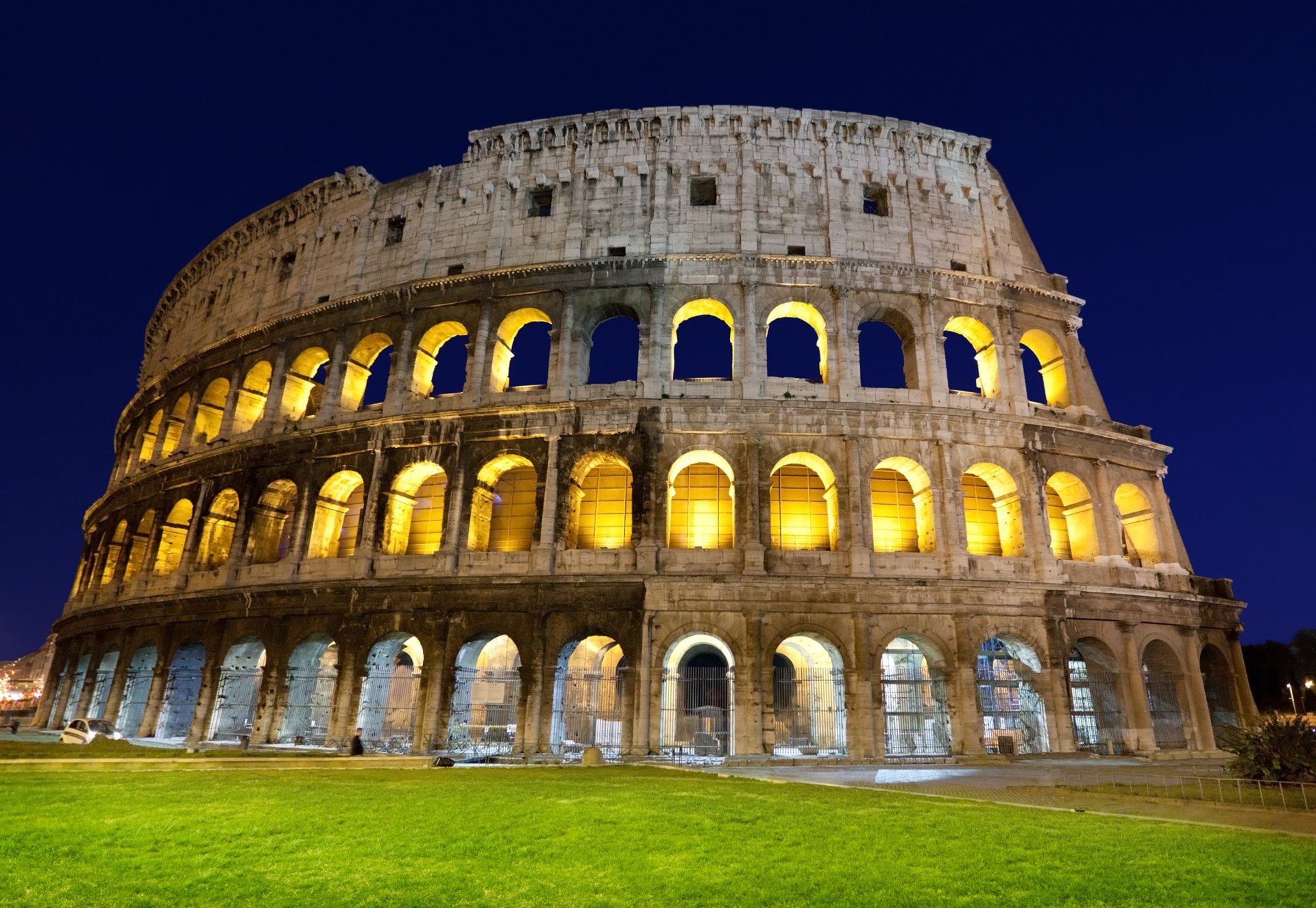 The illuminated Colosseum, a man-made ancient Roman amphitheater, stands majestically against a deep blue night sky.