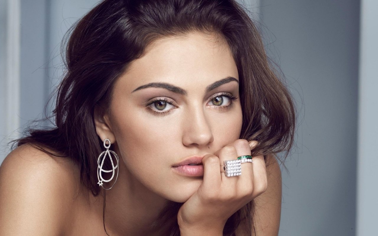 Close-up portrait of celebrity Phoebe Tonkin with wavy brown hair, wearing elegant earrings and a sparkling ring, resting her chin on her hand against a neutral background.