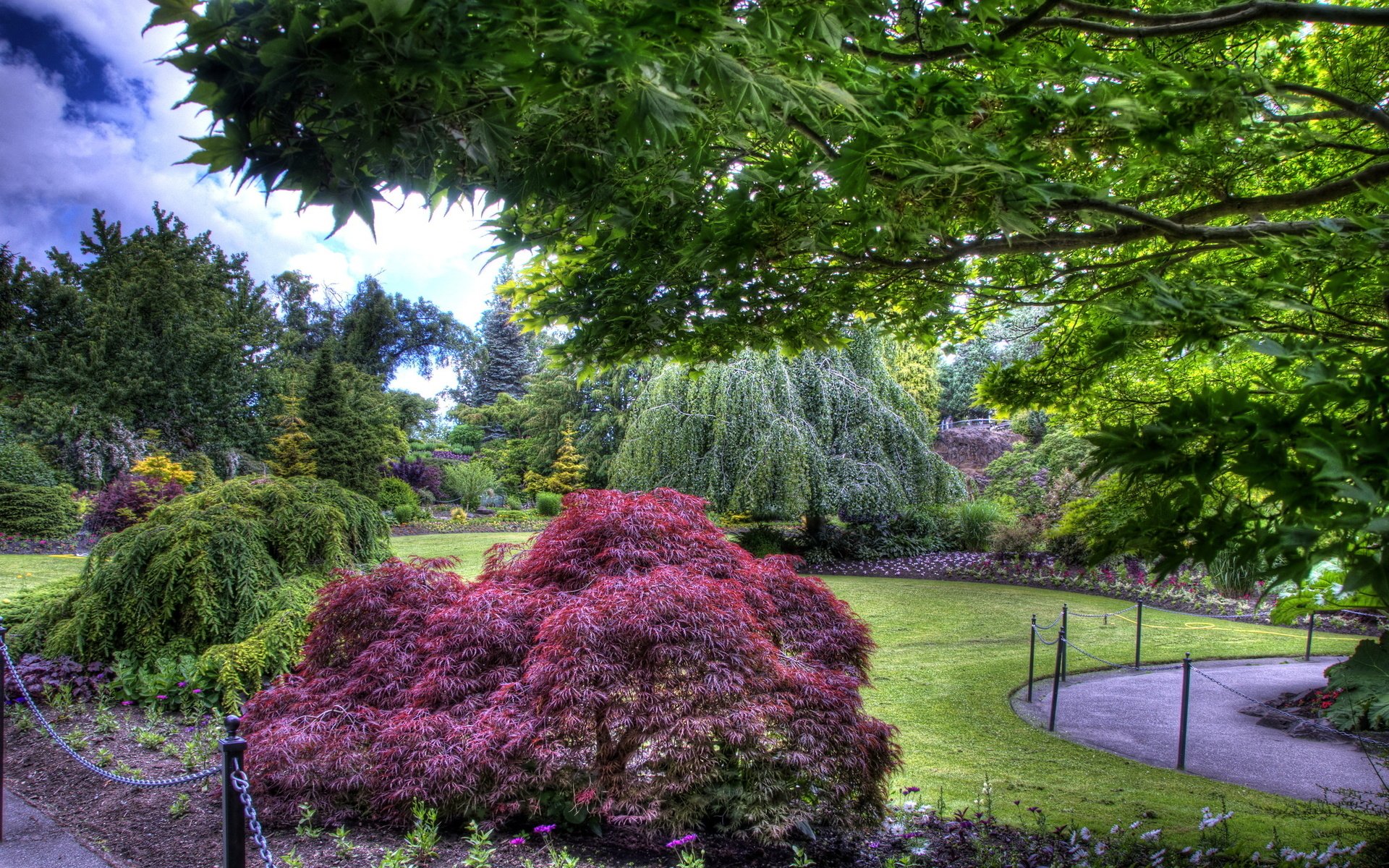 Lush greenery and vibrant shrubs fill this park, showcasing a serene landscape with trees and well-manicured lawns under a partly cloudy sky.