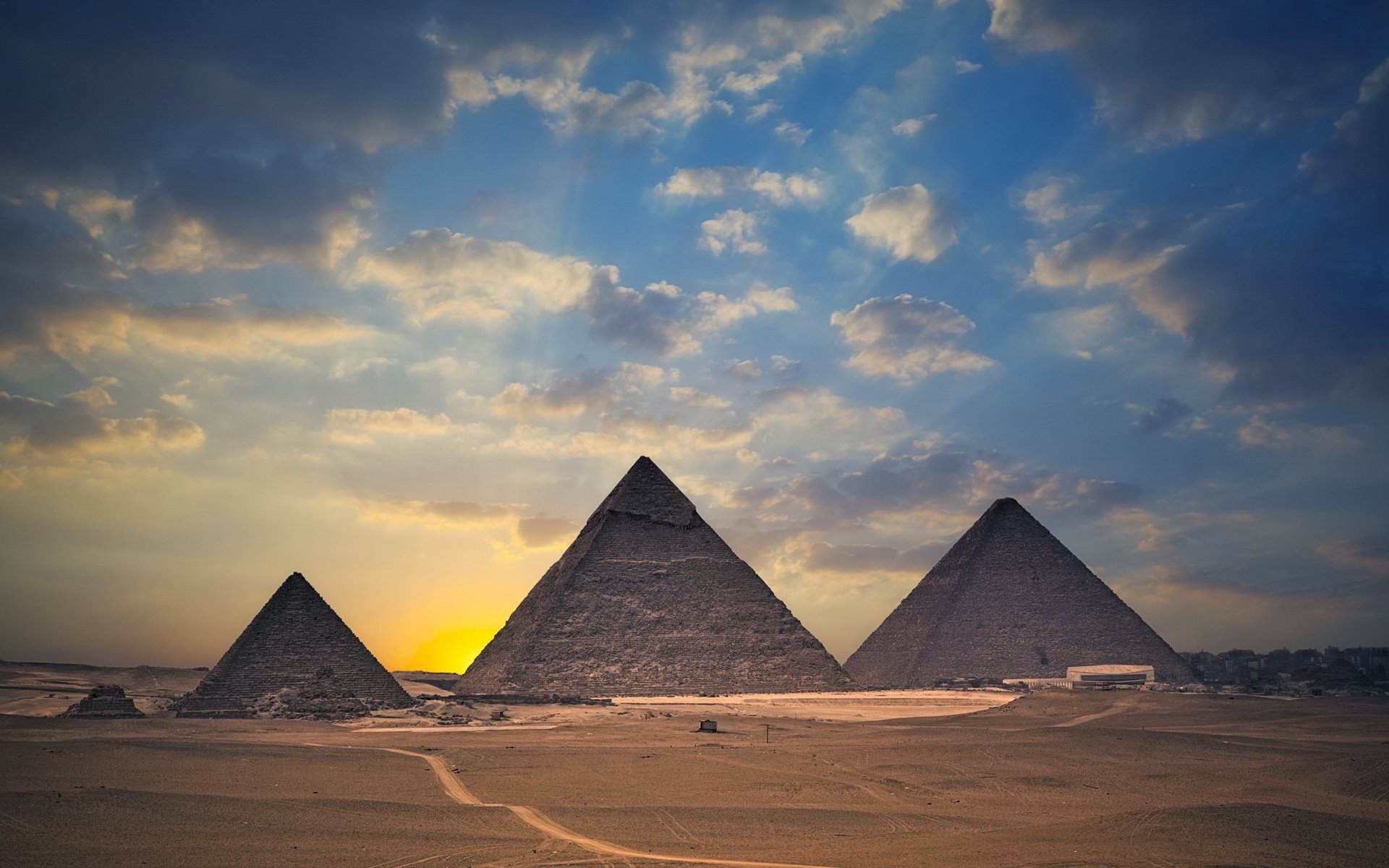 Three man-made pyramids stand on a vast desert plain at sunset beneath a dramatic, cloud-streaked sky.