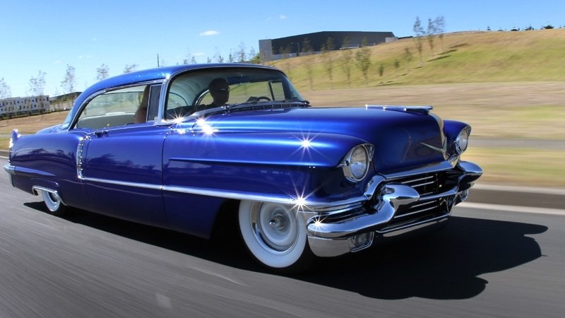 Glossy blue 1956 Cadillac cruising on a highway, low stance with shining chrome bumpers and whitewall tires.