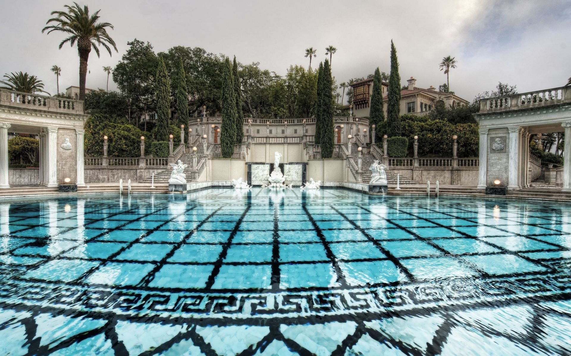 A serene man-made pool with clear blue water, surrounded by ornate architecture, statues, and tall palm trees under a cloudy sky.