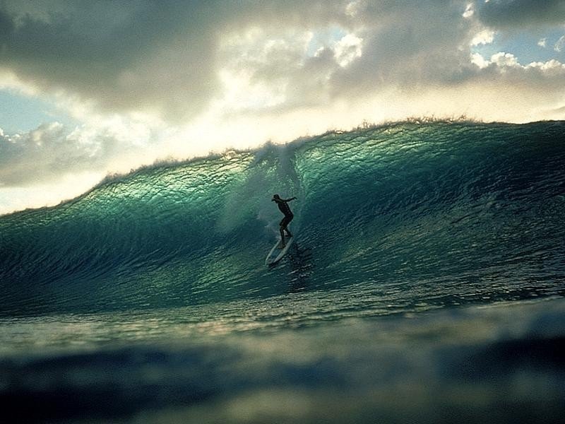 A surfer skillfully rides a large wave, showcasing the dynamic energy of the sport against a backdrop of dramatic clouds and shimmering water.
