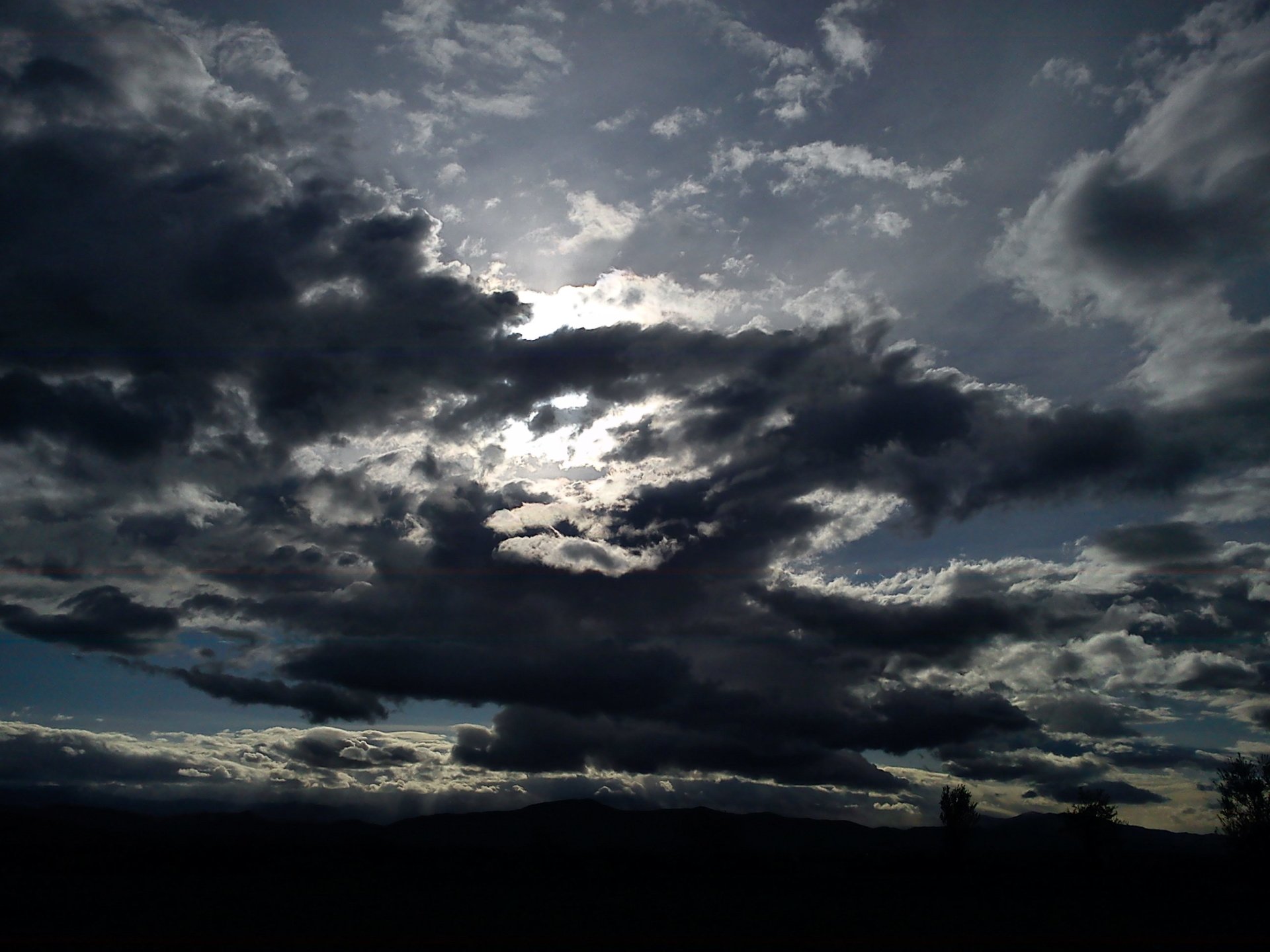 Dark clouds gather over a natural landscape, with sunlight breaking through gaps, creating a dramatic sky scene.