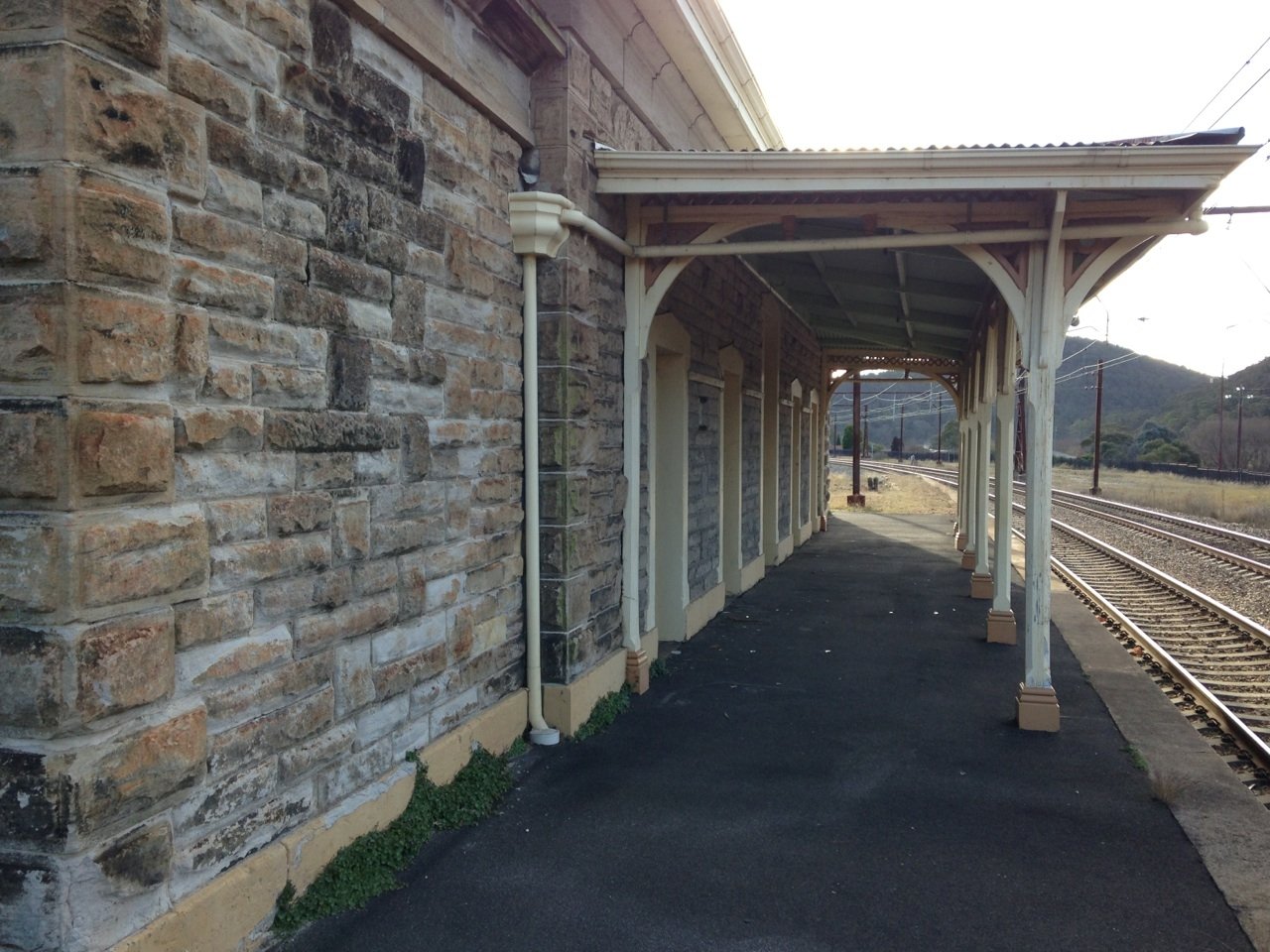 Stone building with a covered platform next to railroad tracks at a train station in a rural area.