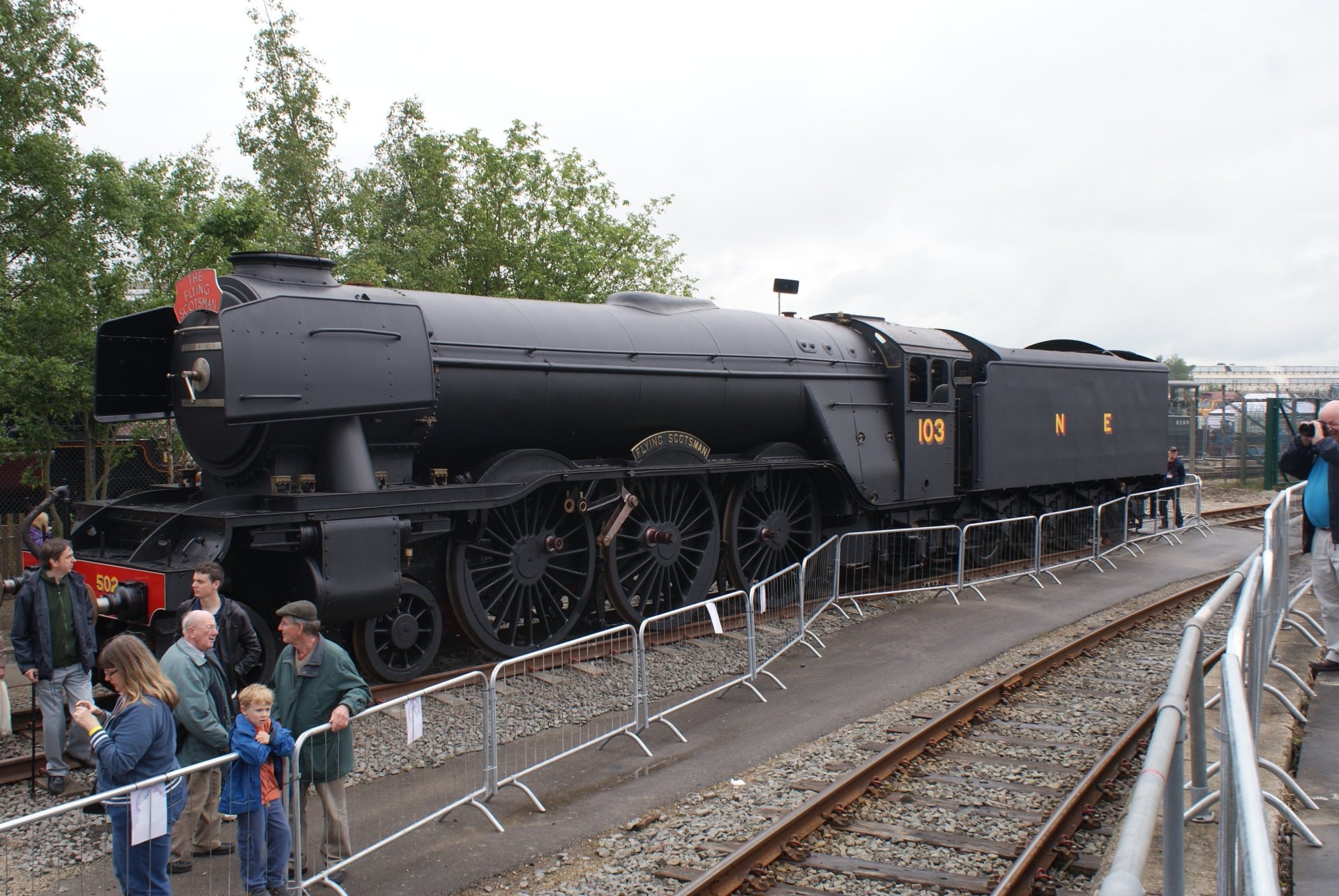 A vintage black steam train vehicle displayed outdoors with people observing behind barriers along the railway tracks.