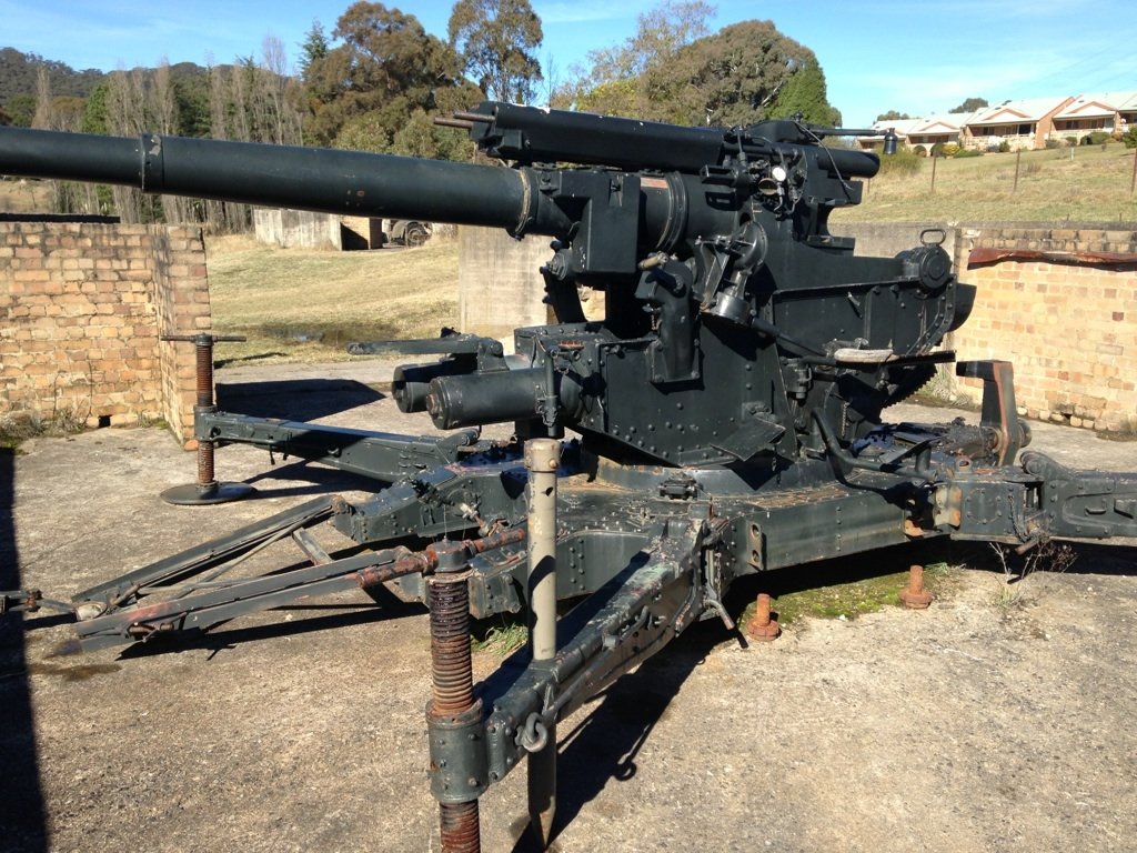 A World War II-era military artillery gun displayed outdoors, showcasing detailed metal components and a long barrel against a backdrop of trees and rustic buildings.
