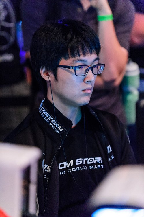 A focused esports gamer competes in a Starcraft video game competition, wearing branded apparel and earphones amid a crowded event setting.