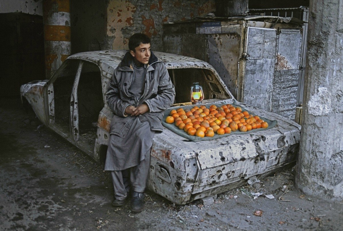 A man sits beside a street vendor display of fruit arranged on the hood of an abandoned, rusted car inside a dilapidated building.