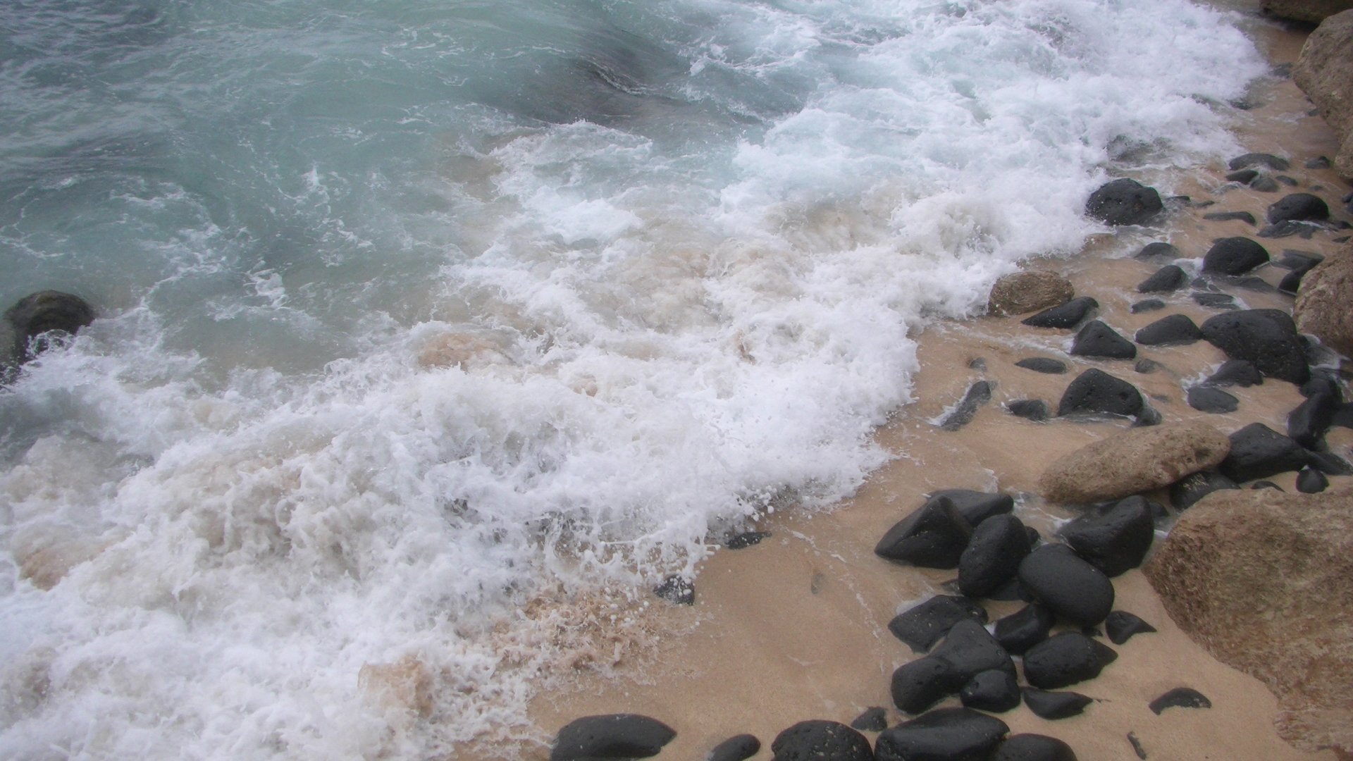 Waves crash onto a sandy beach scattered with dark rocks, showcasing the natural beauty of water meeting the shore.