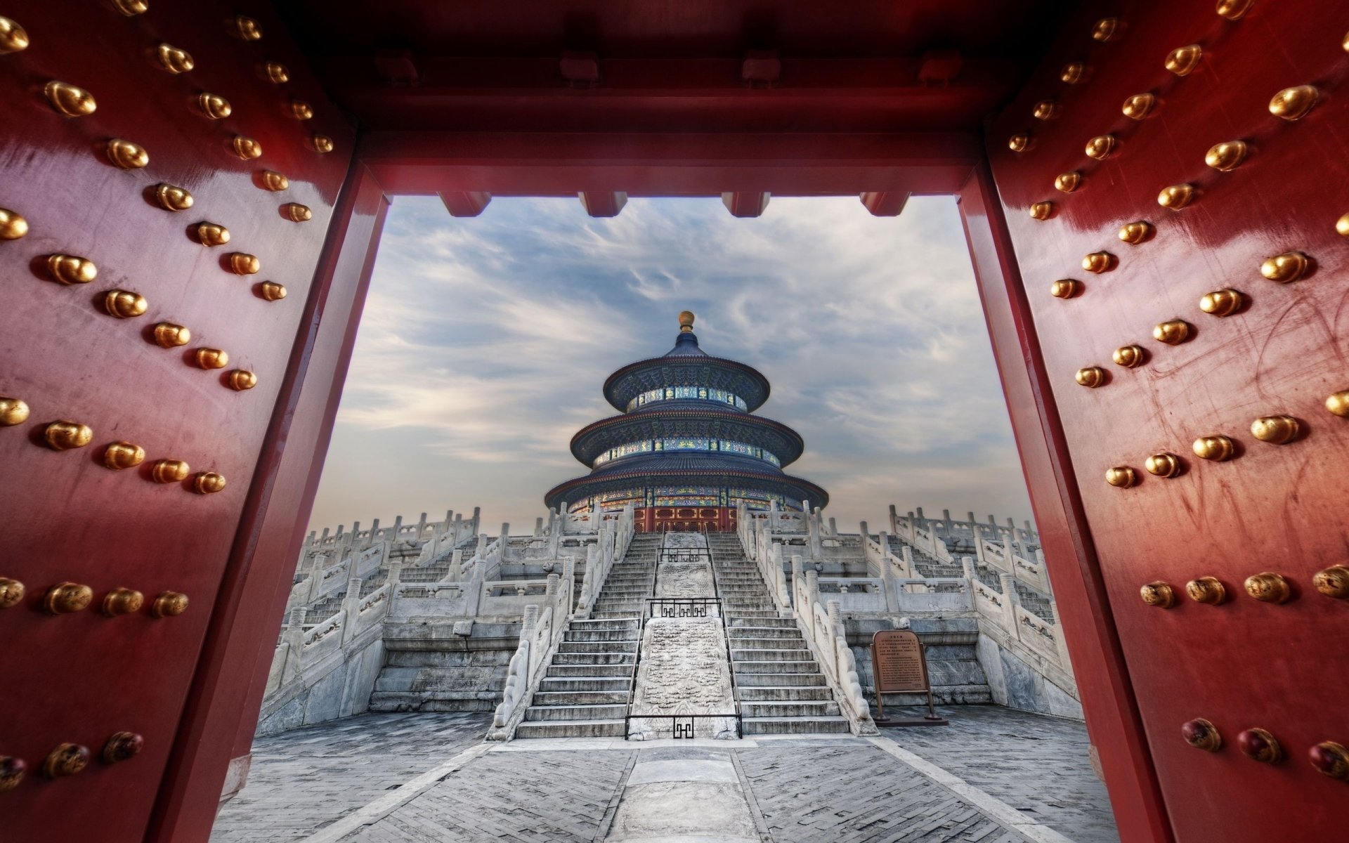 View of the Temple of Heaven framed by a large red gate with golden studs, showcasing its religious and architectural significance under a cloudy sky.