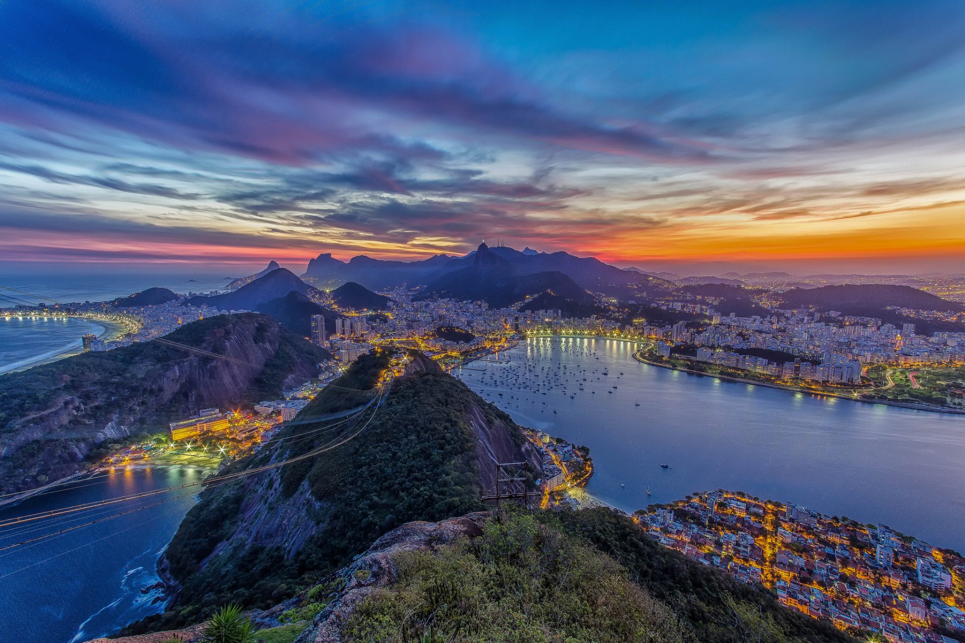 A breathtaking view of Rio de Janeiro at sunset, showcasing the city's skyline and dramatic landscapes along the coastline and hills, illuminated by evening lights.