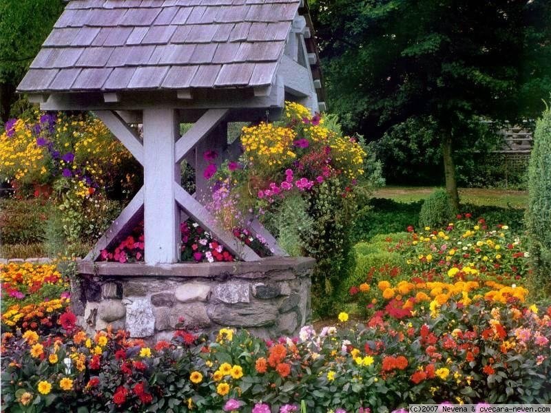 A man-made stone well with a wooden roof surrounded by vibrant flowers in a lush garden setting.