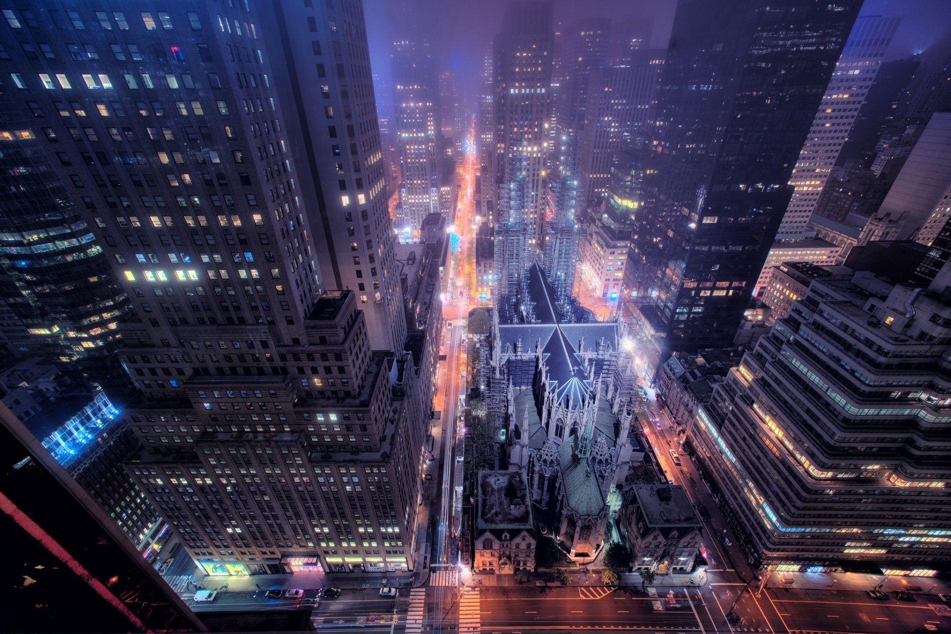 Aerial night view of the religious landmark St. Patrick's Cathedral nestled among illuminated skyscrapers, its Gothic spires and ornate façade glowing amid city lights.