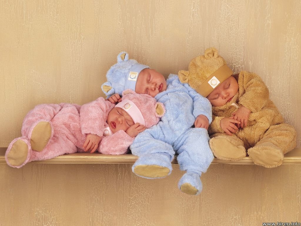 Three sleeping babies dressed in colorful bear-themed outfits lie peacefully on a shelf against a soft, neutral background, capturing a moment of serene childhood innocence.