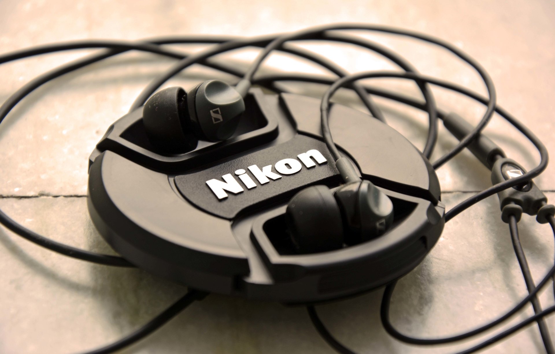 Close-up of a man-made Nikon lens cap tangled with wired in-ear headphones resting on a tiled surface.