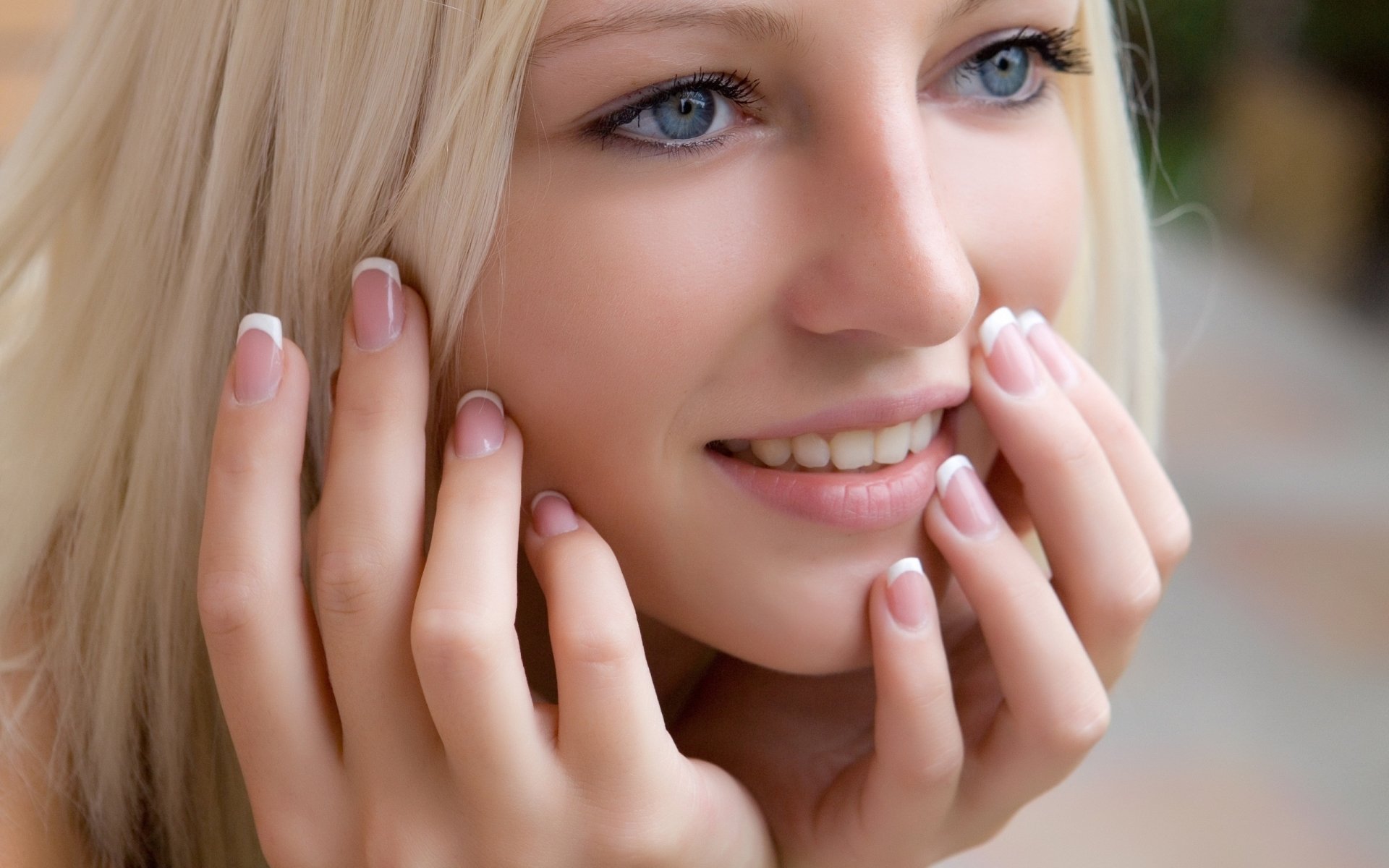 Close-up of a smiling blonde woman touching her face, showing blue eyes and manicured nails.