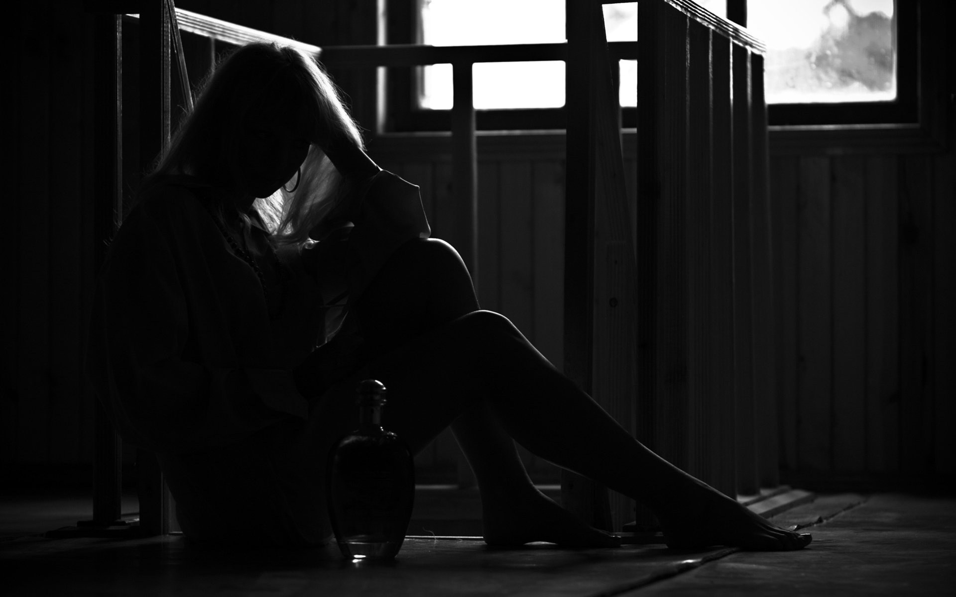 Silhouetted woman seated on a wooden floor by a window, head resting on her hand, a bottle beside her.