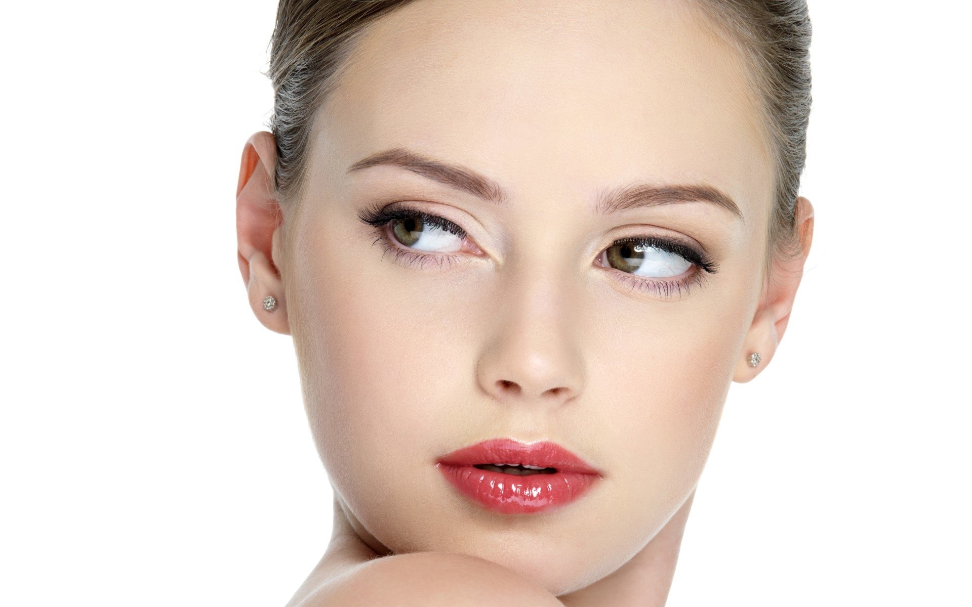 Close-up portrait of a young woman model with glossy red lips, flawless skin and subtle eye makeup, gazing to the side against a white background.