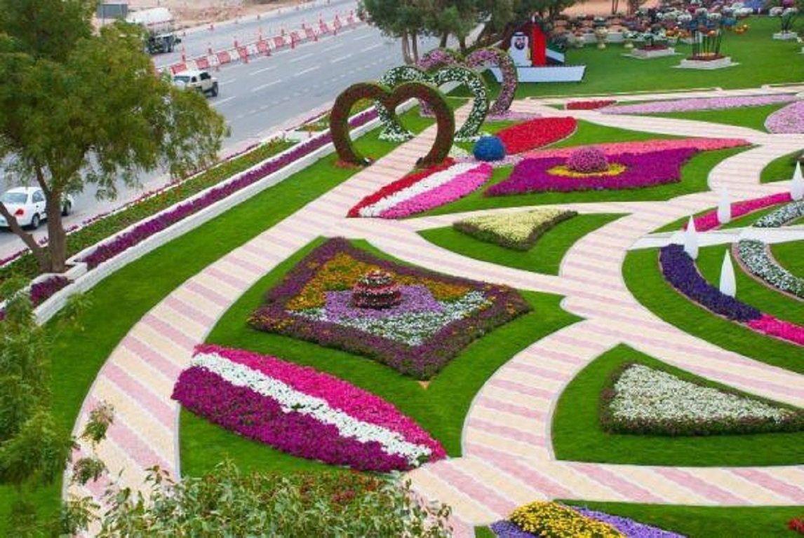 A colorful man-made garden featuring vibrant flower patches and winding pathways creates an inviting outdoor space, surrounded by greenery and a bustling road in the background.
