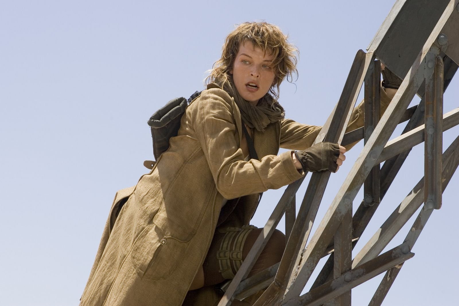 Milla Jovovich as Alice from Resident Evil: Extinction climbs a metal ladder against a clear sky, dressed in a long coat and tactical gear.