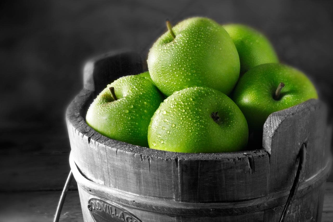 A rustic wooden basket holds a collection of fresh green apples, glistening with water droplets, set against a black and white background.