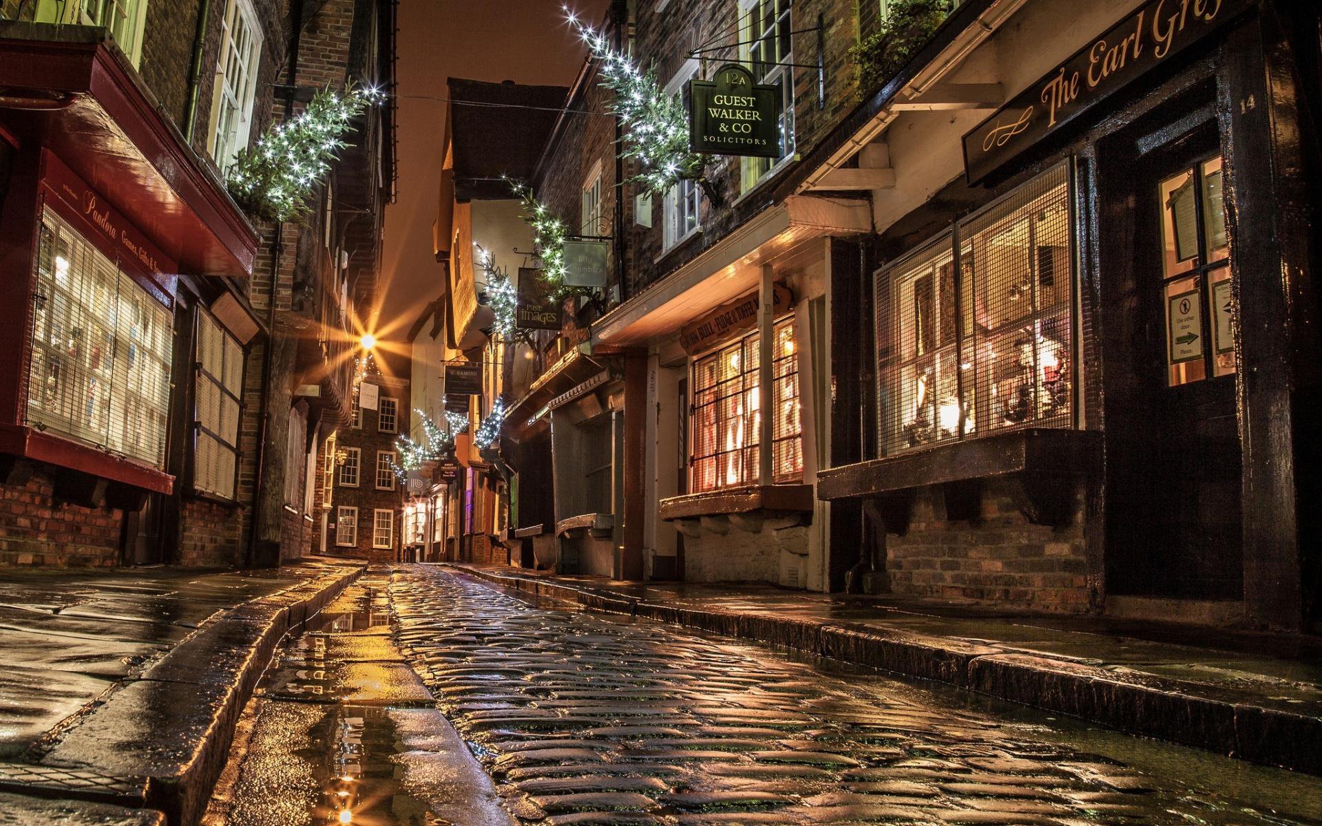 A quiet, man-made cobblestone street in a village at night, illuminated by warm lights from shop windows and festive string lights overhead.