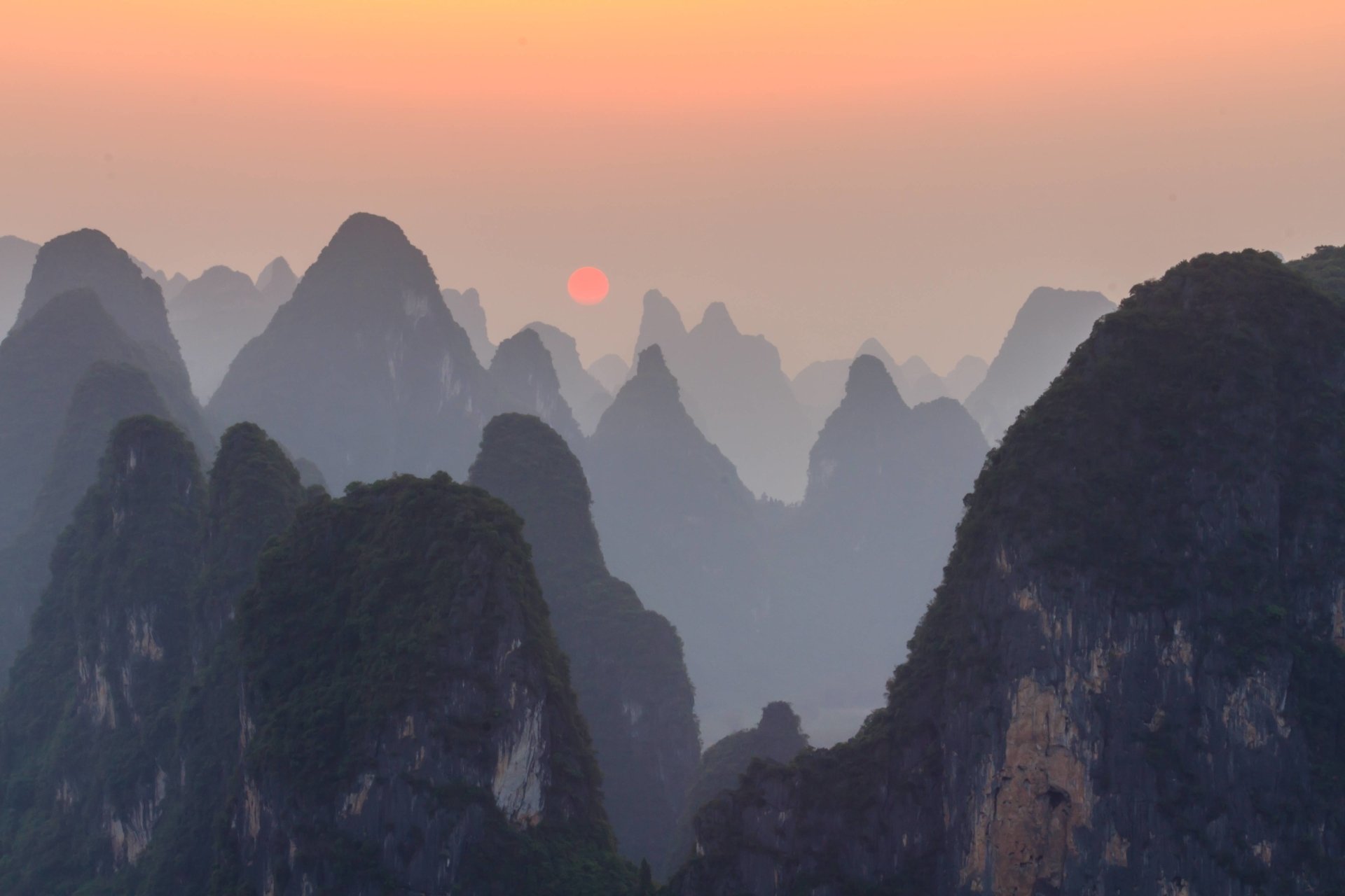 Sun sets behind foggy mountain peaks, casting a soft glow over the serene natural landscape.