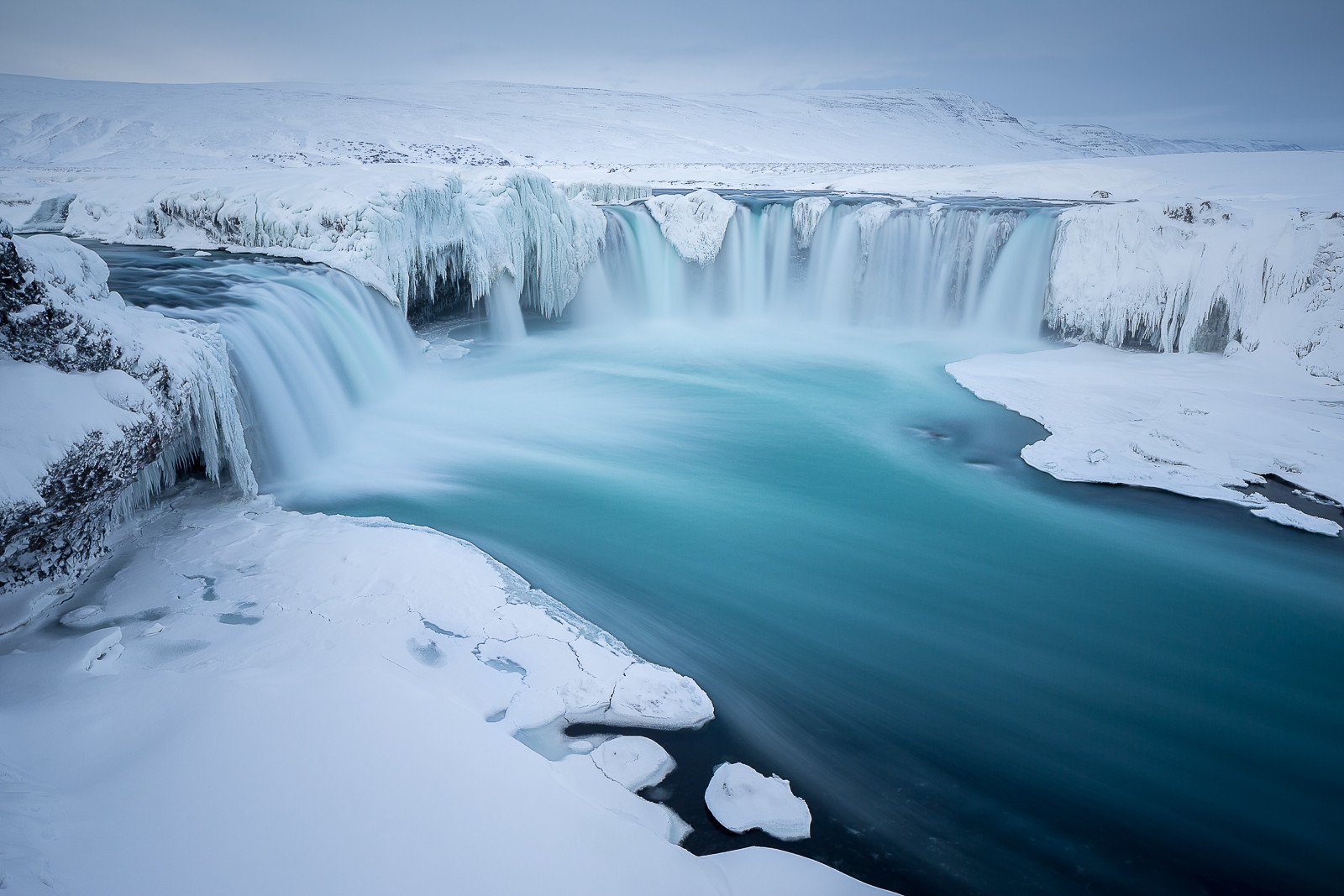 A serene ice-covered landscape featuring a striking waterfall cascading into a turquoise lake, surrounded by snowy terrain in a tranquil natural setting.
