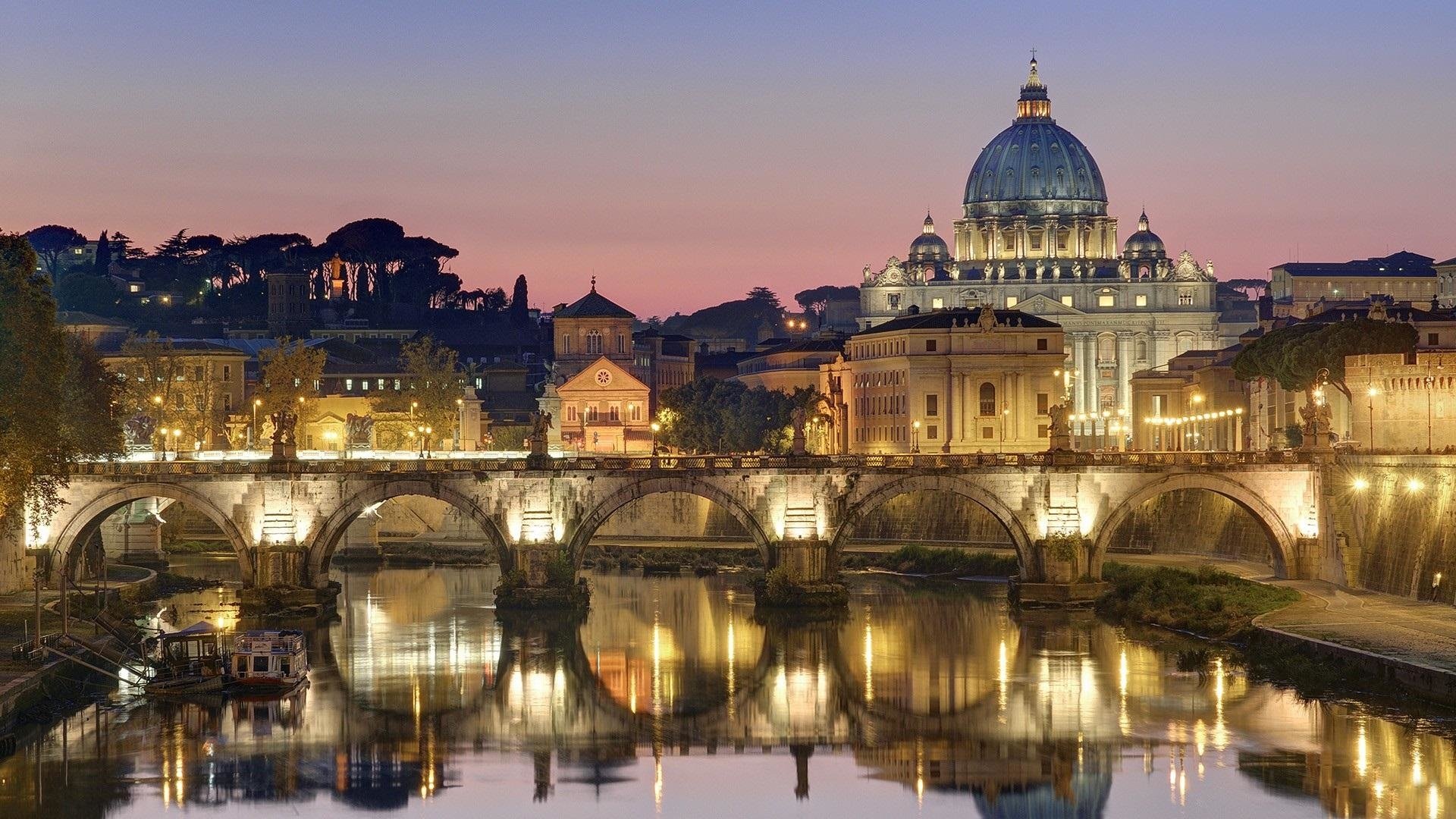 Dusk man-made cityscape with an illuminated domed cathedral and arched stone bridge reflected in a calm river.
