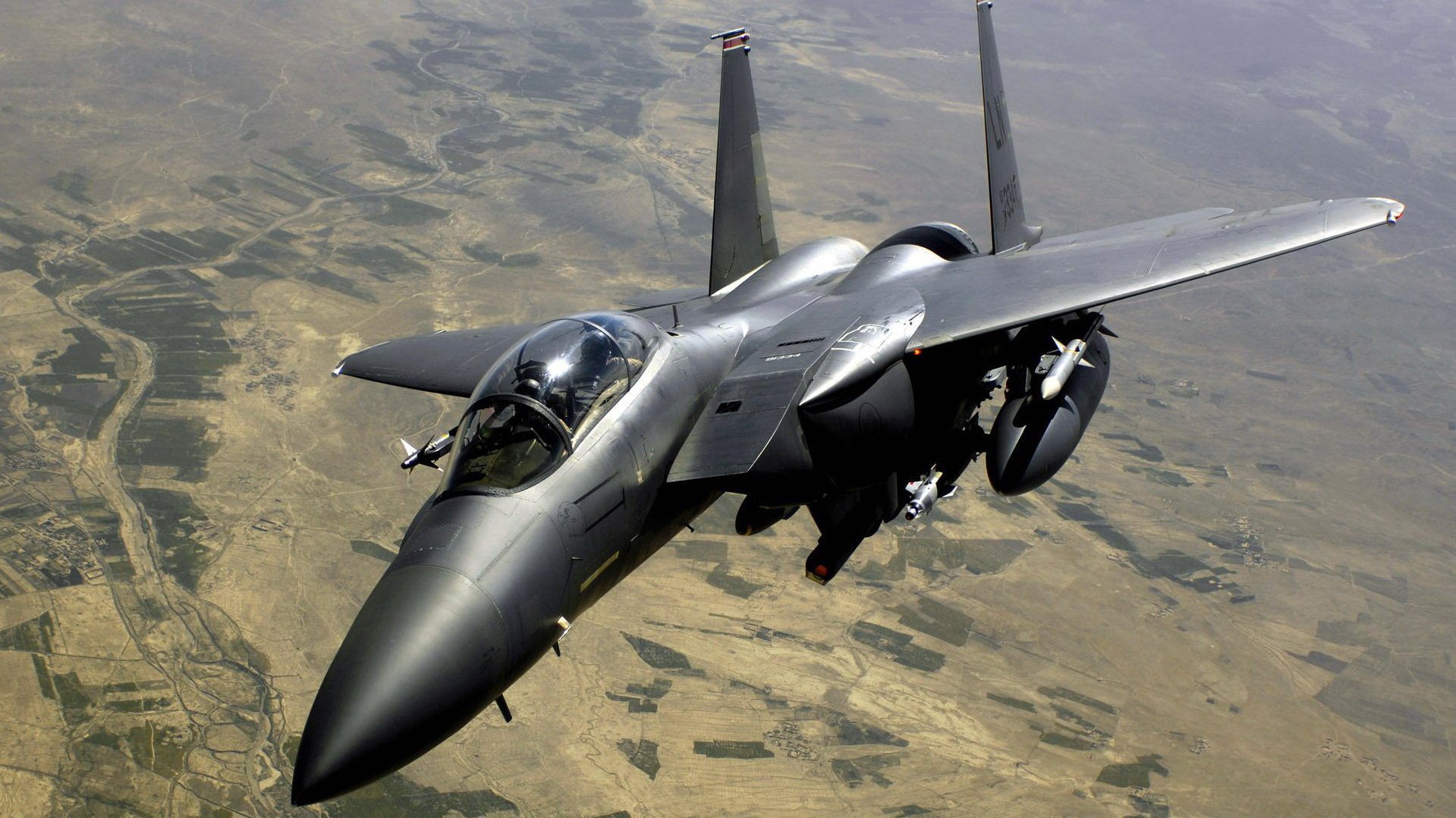 McDonnell Douglas F-15 Eagle military fighter jet flying low over arid terrain, seen from a front-left aerial perspective.
