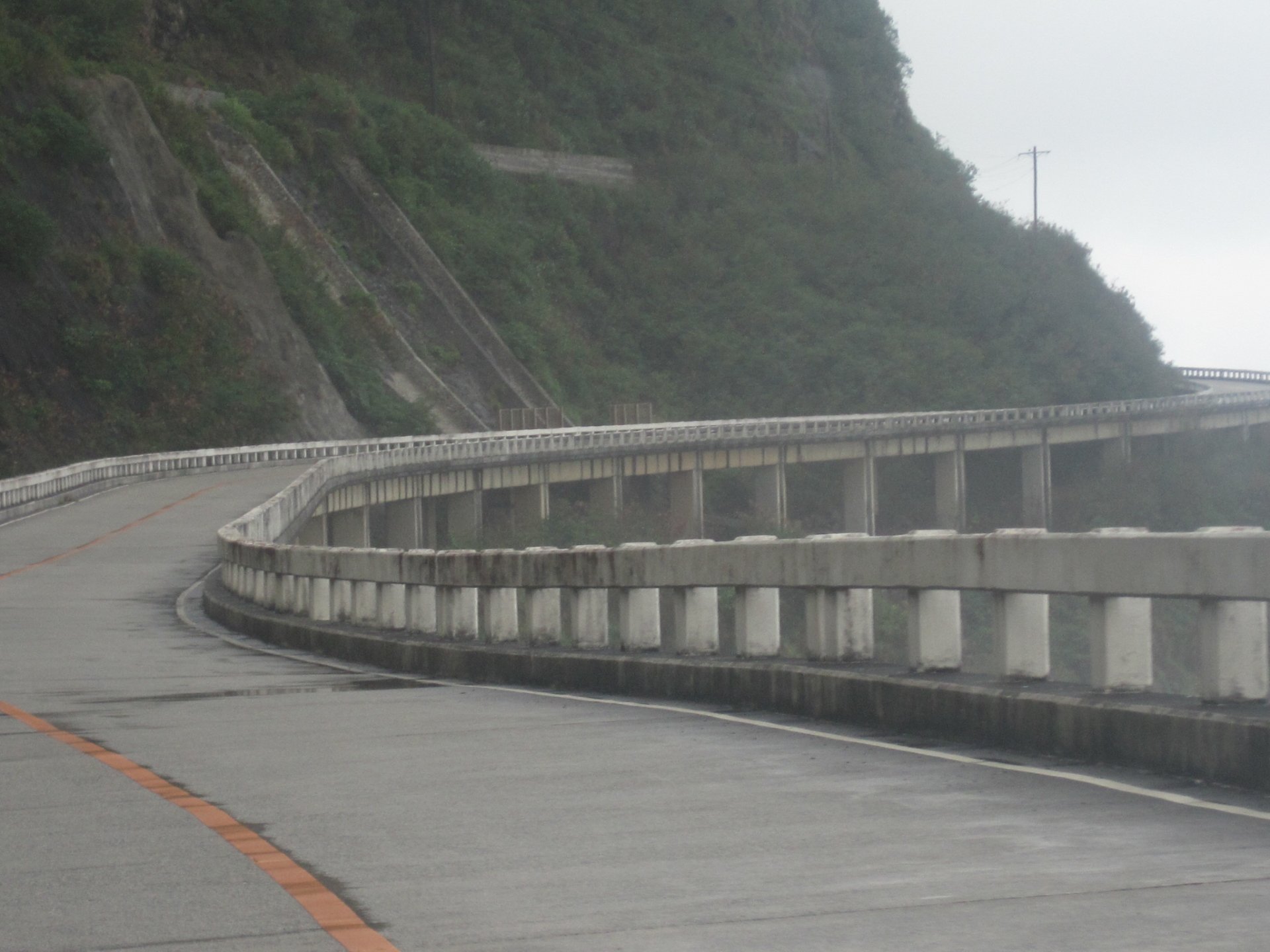 A man-made concrete road curves along a rocky hillside with guardrails on the outer edge and a misty atmosphere in the background.