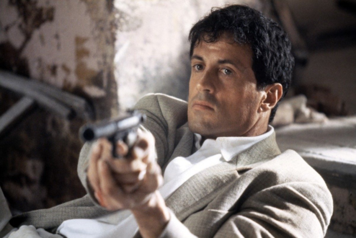 Still from the movie Assassin (1995): a suited man reclines amid rubble, pointing a handgun straight toward the viewer.