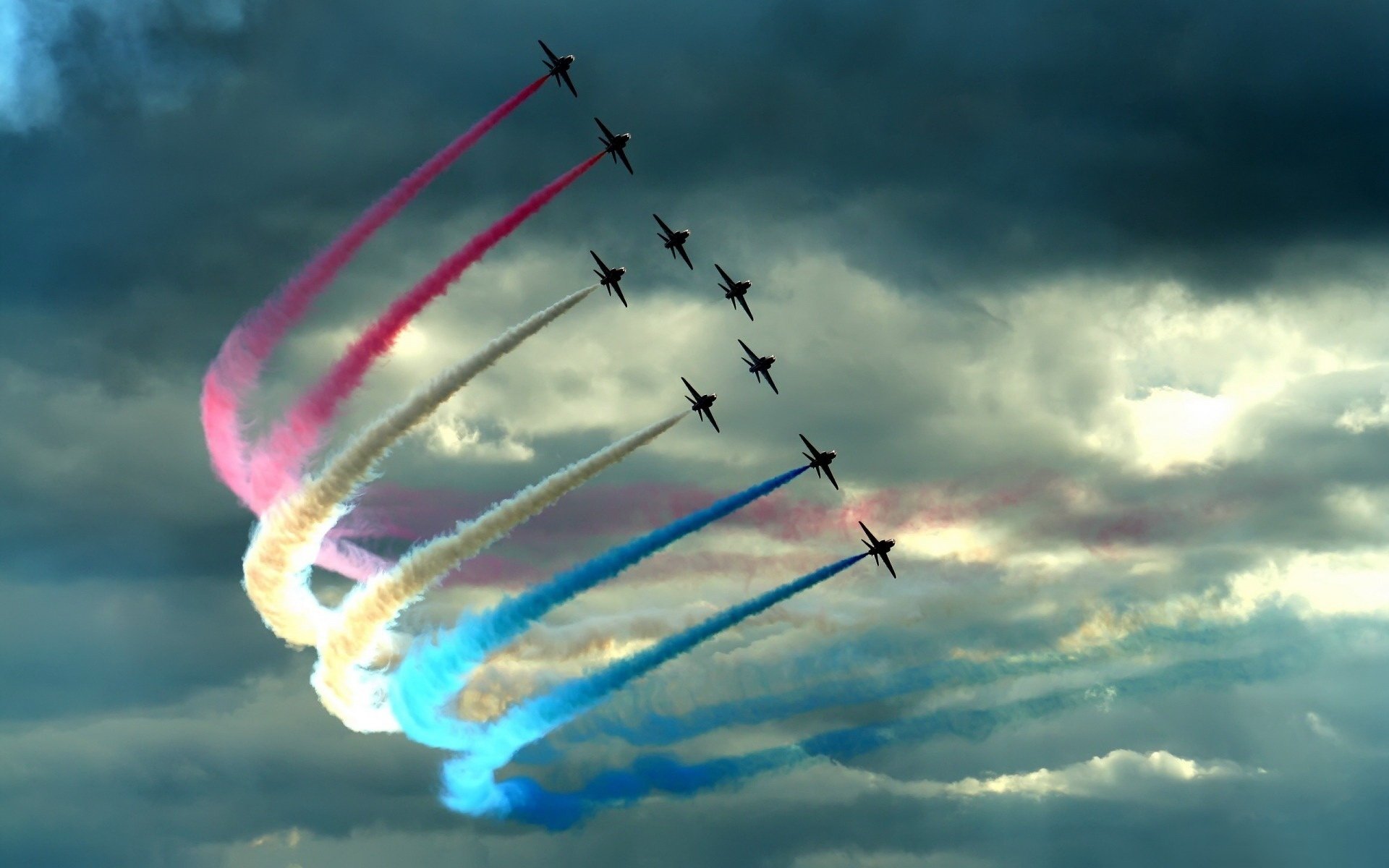 Military jets in tight formation at an air show sweep through a cloudy sky, leaving red, white and blue smoke trails.