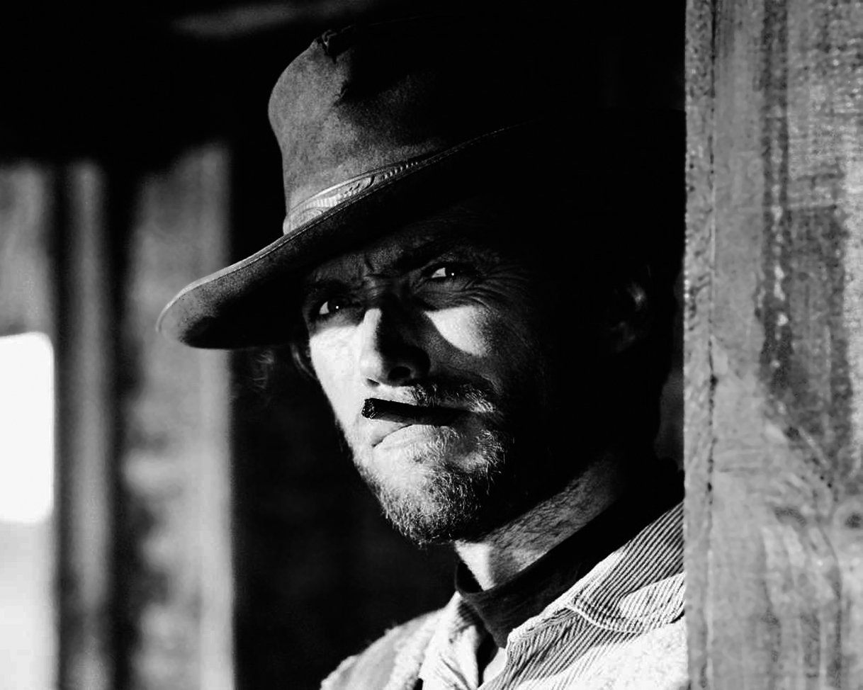 A black and white portrait of Clint Eastwood, showcasing his rugged demeanor with a hat and cigarette, standing in a doorway that enhances his intense expression.