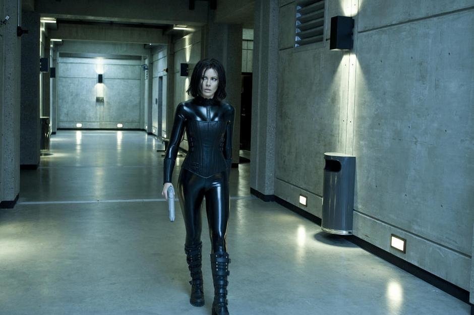 A character in a black leather suit holding a gun walks confidently through a dimly lit corridor in the movie Underworld: Awakening.