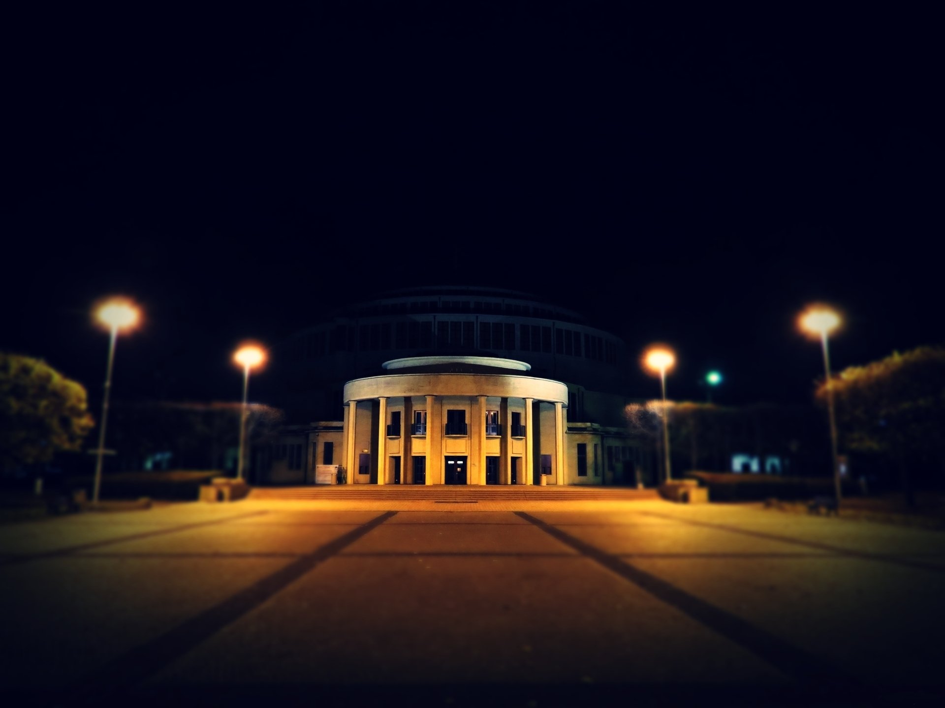 A lit building at night with a prominent circular entrance, surrounded by landscaped paths and lamp posts, showcasing an intriguing man-made architectural design.