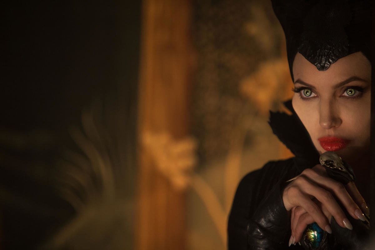 Angelina Jolie as Maleficent, with striking makeup and dark attire, gazes intensely in a dimly lit, mystical setting.