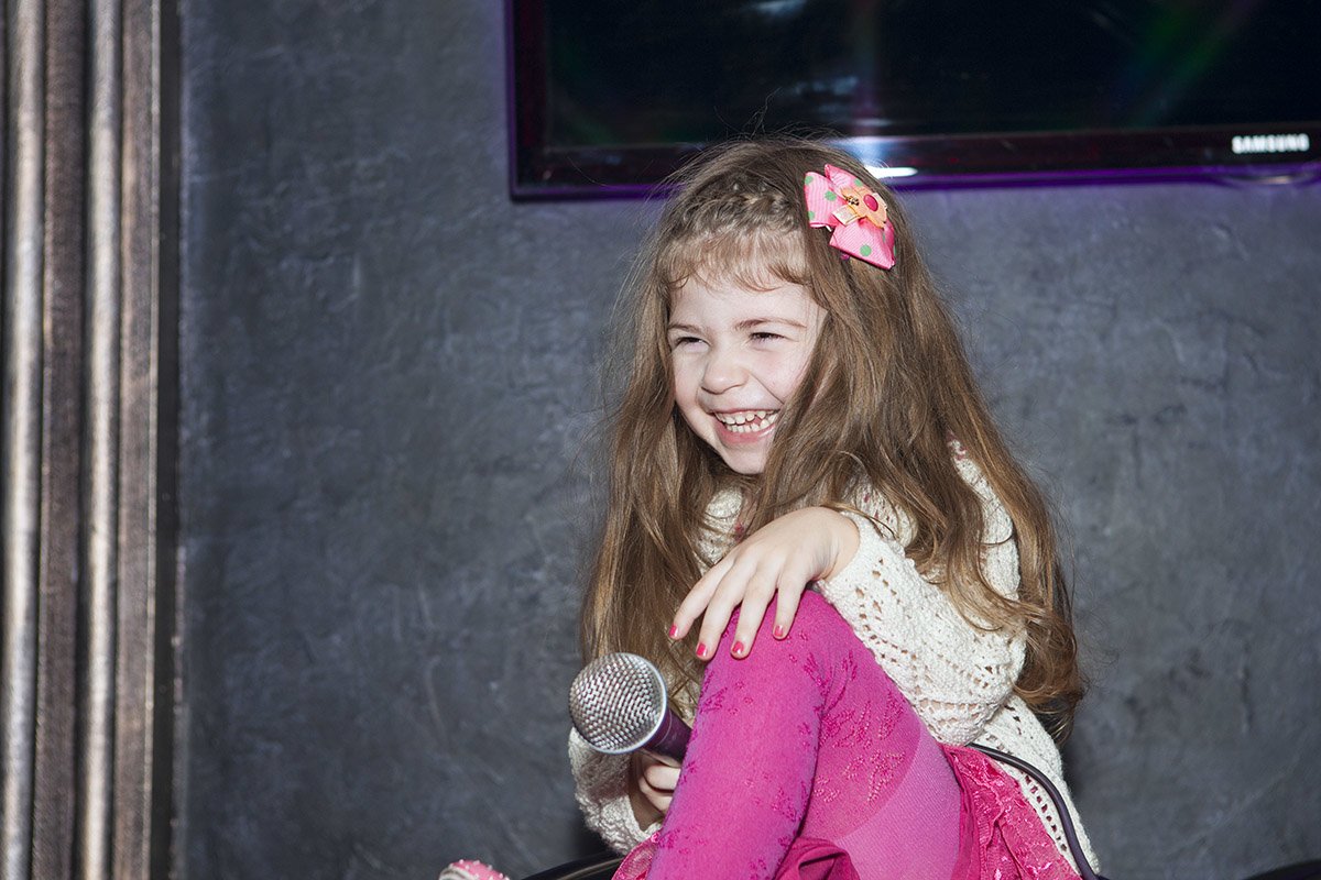 A joyful child with long hair and a flower hair accessory sits with a microphone, laughing heartily in a cozy indoor setting. The image captures her playful spirit beautifully.
