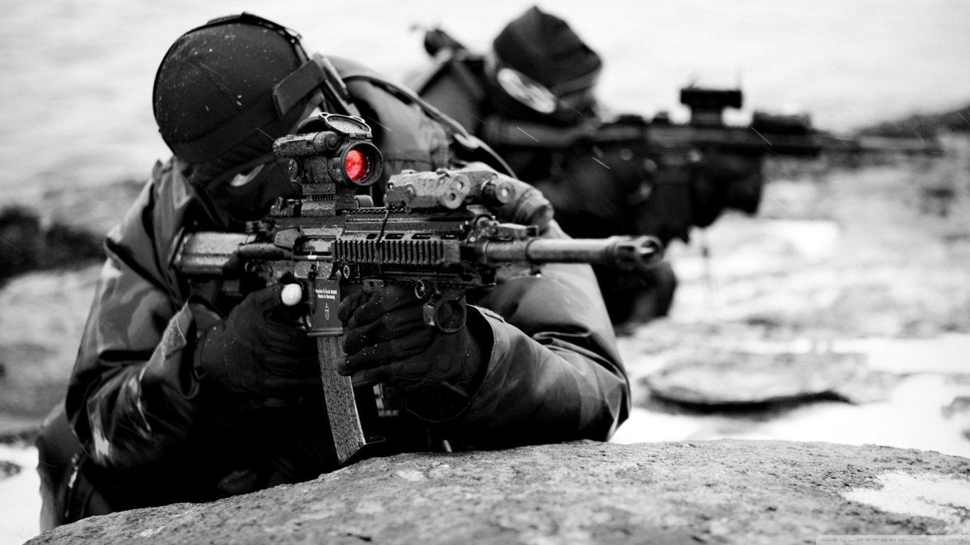 A military sniper, dressed in tactical gear, aims a rifle with a red dot sight, while another soldier provides support in the background. The scene captures a tense, focused moment.
