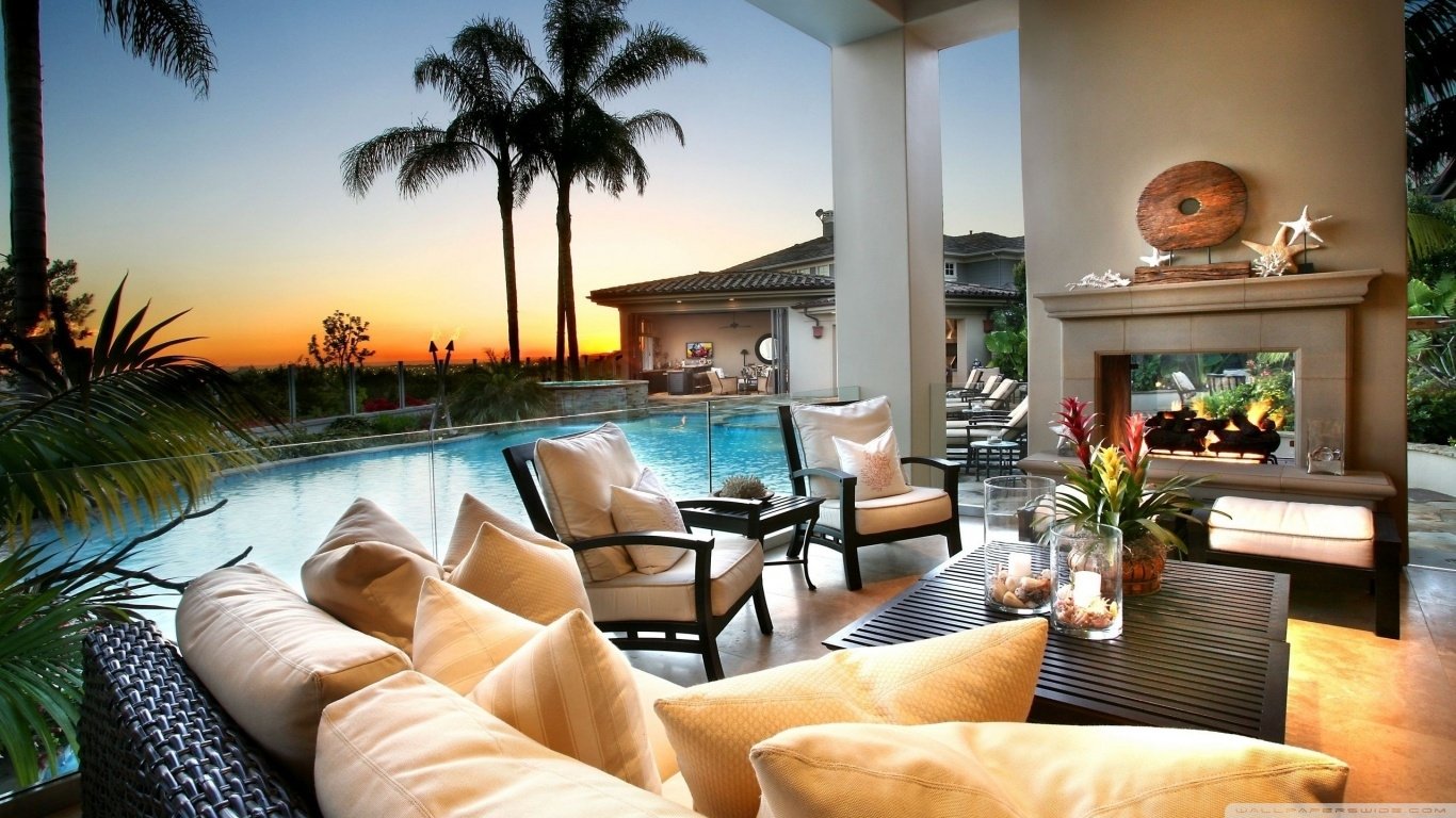 A man-made outdoor room with cushioned seating, a fireplace, and tropical plants overlooking a pool at sunset.