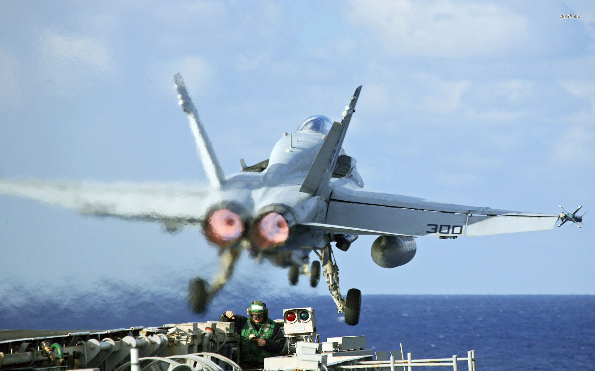 A McDonnell Douglas F/A-18 Hornet takes off from an aircraft carrier, with powerful exhaust flames and a crew member visible on deck, against a backdrop of blue ocean and sky.