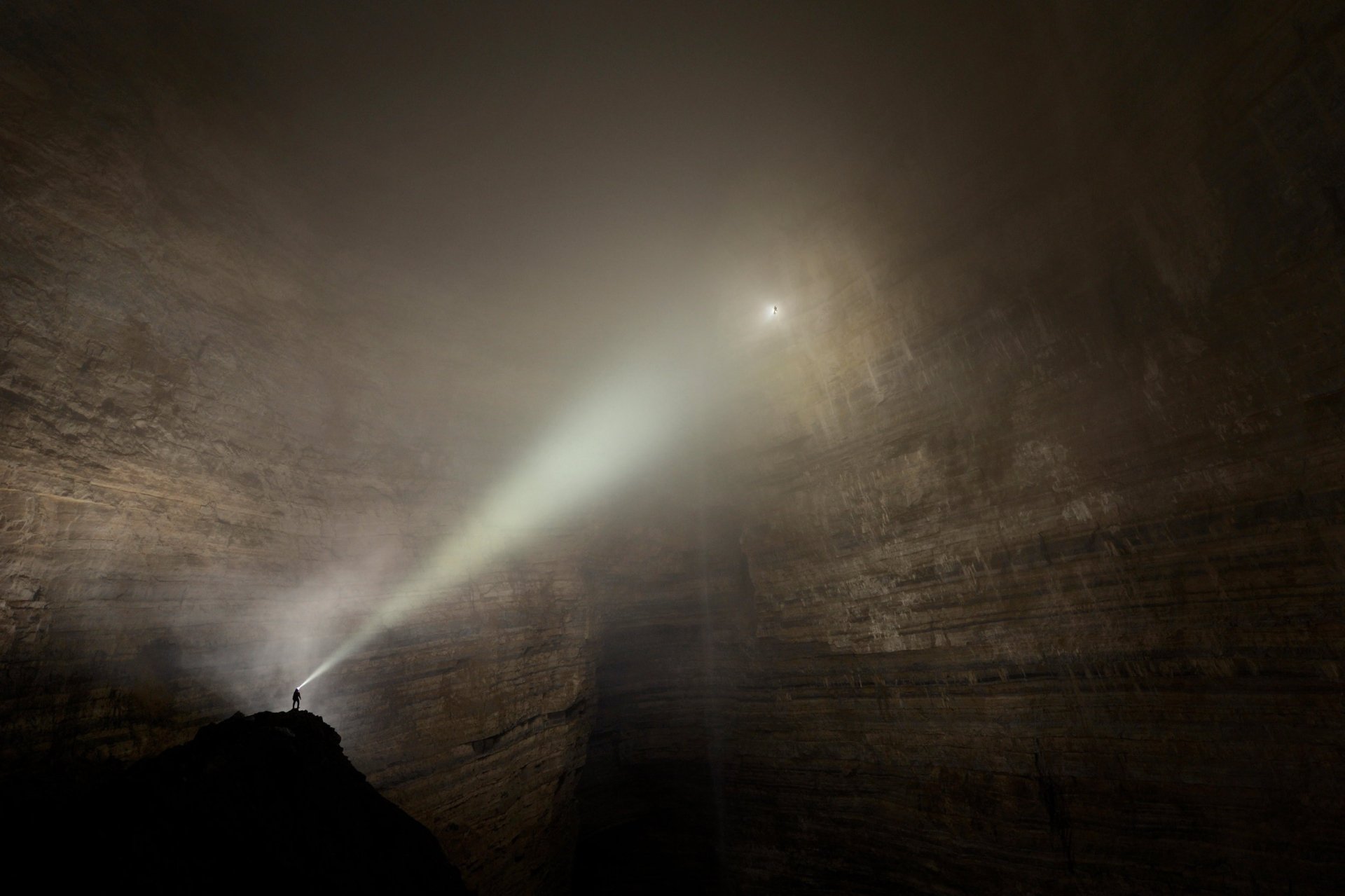 An explorer in a vast natural cave shines a beam of light through mist, capturing the thrill of cave exploration and adventure.
