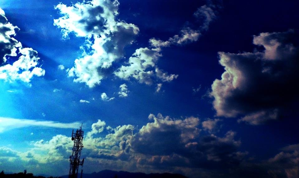 A vibrant nature scene showcasing a deep blue sky filled with scattered clouds above a silhouette of mountains and a communication tower.