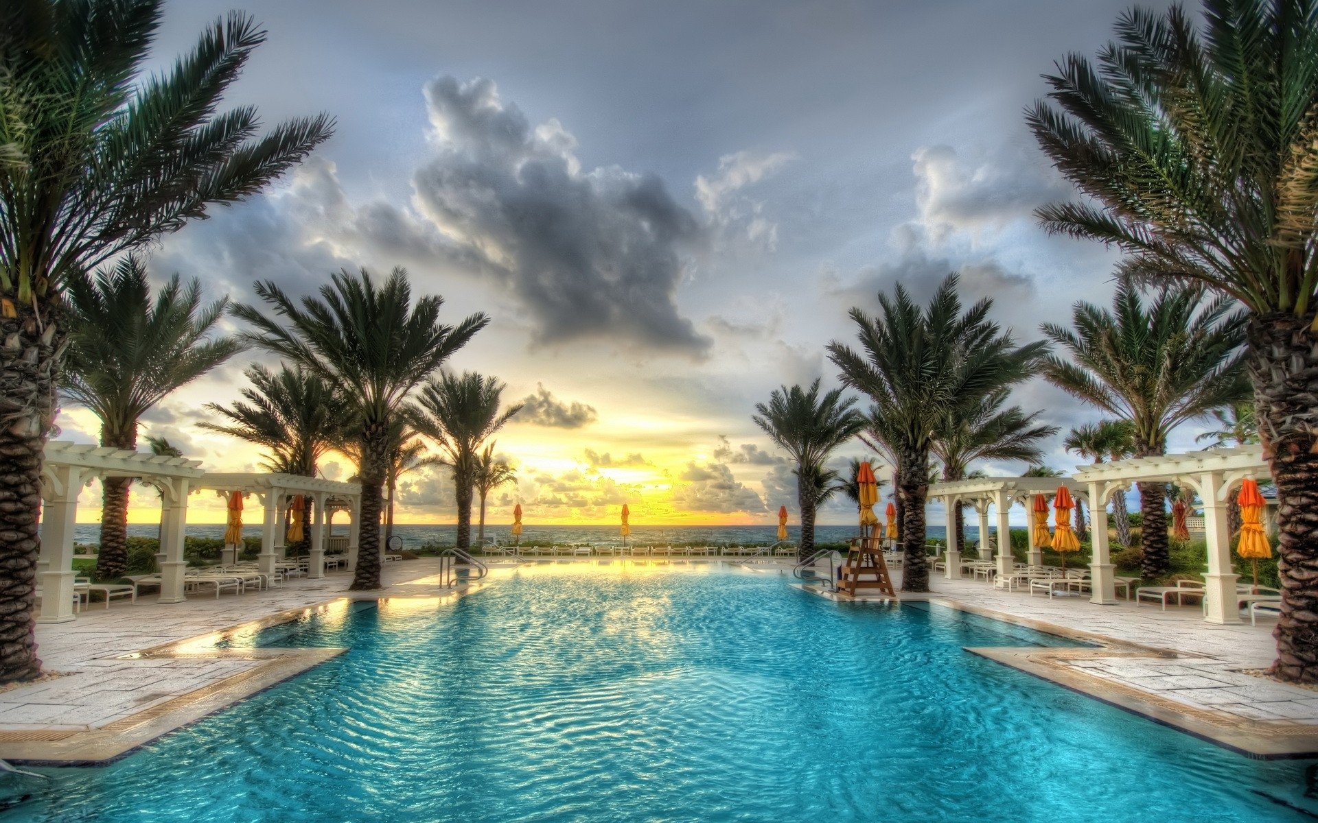 A man-made tropical pool framed by palm trees and white cabanas, with a dramatic sunset sky over the horizon.