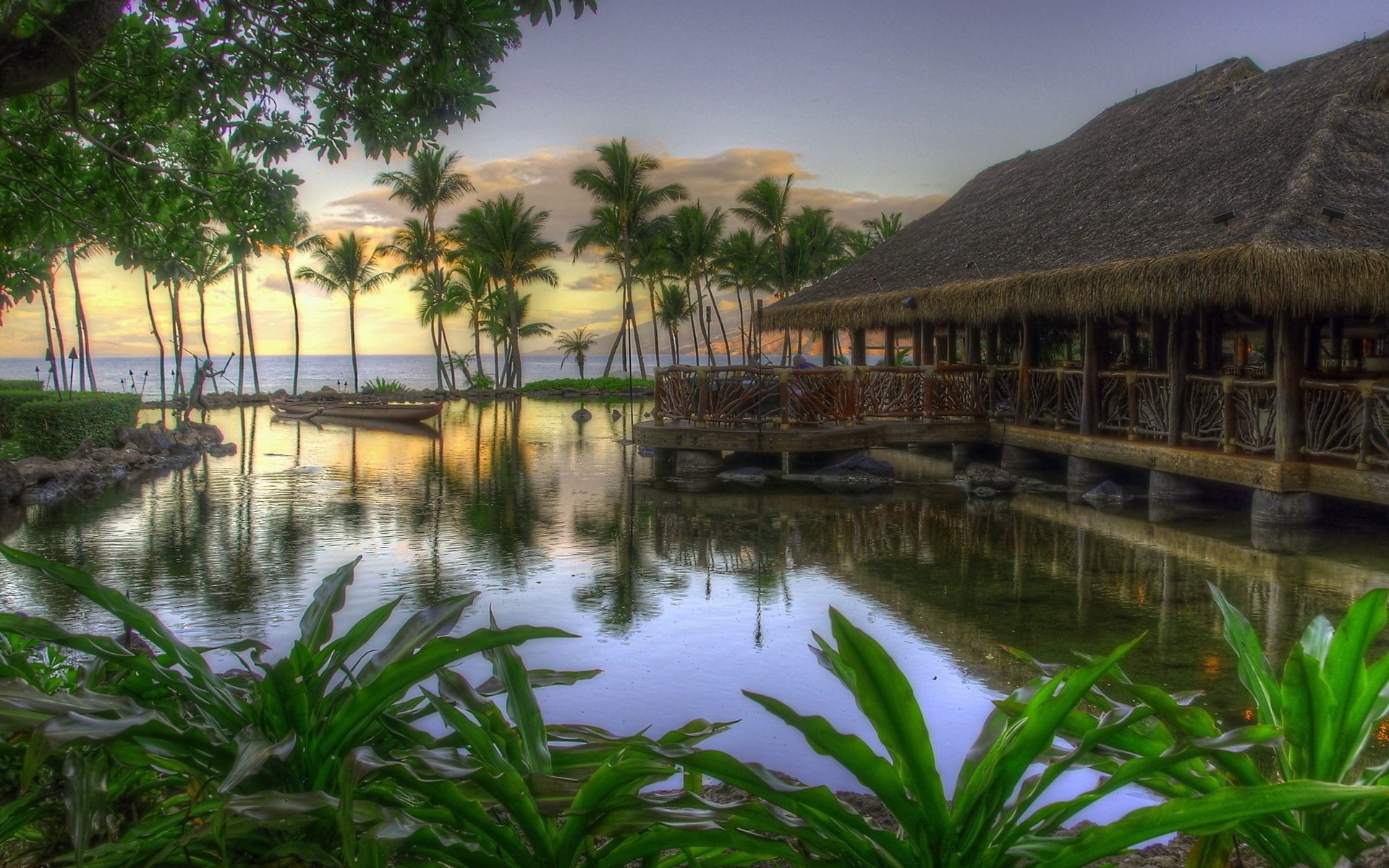 A serene resort with thatched bungalows beside a calm pond, surrounded by palm trees and lush greenery, overlooking the ocean at sunset.