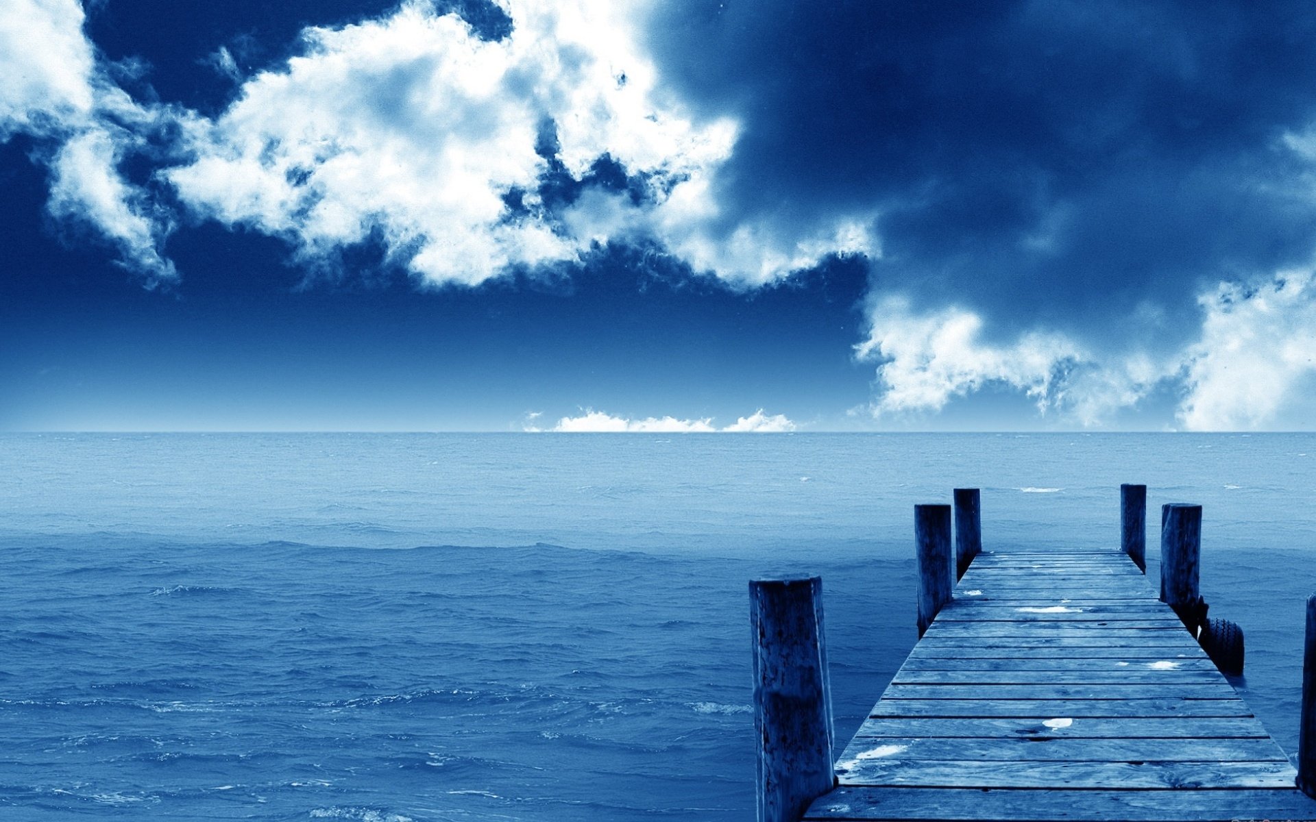 A wooden pier extends into a serene blue ocean under a dramatic sky filled with clouds, showcasing the beauty of the man-made dock against the natural seascape.